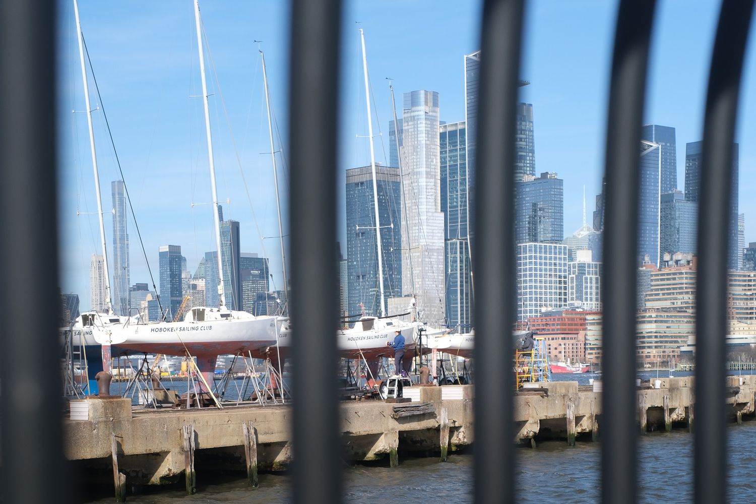 Sailboats docked at a marina with the New York City skyline visible through metal fence railings from Hoboken, NJ.