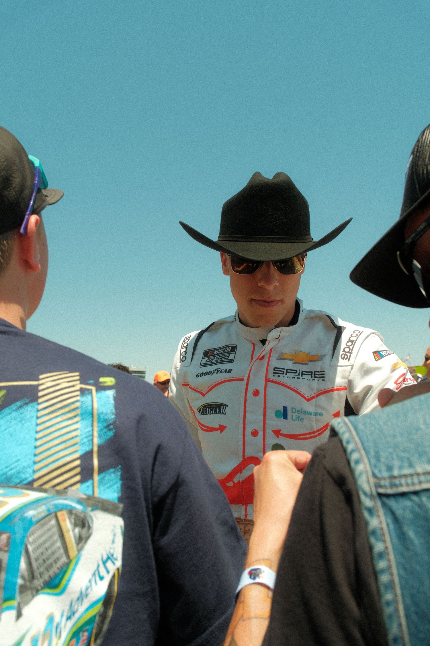 NASCAR driver wearing black cowboy hat and racing suit signing autographs for fans at a race event.