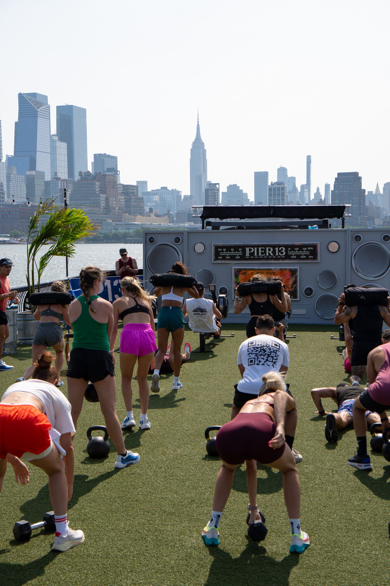 Group fitness class doing kettlebell workouts on turf at Pier 13 with NYC skyline backdrop.