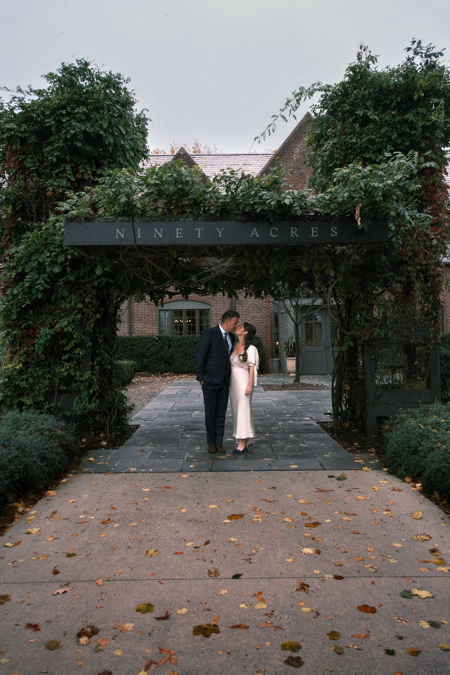 A couple shares a romantic kiss under an ivy-covered arbor at Ninety Acres venue, surrounded by autumn leaves.