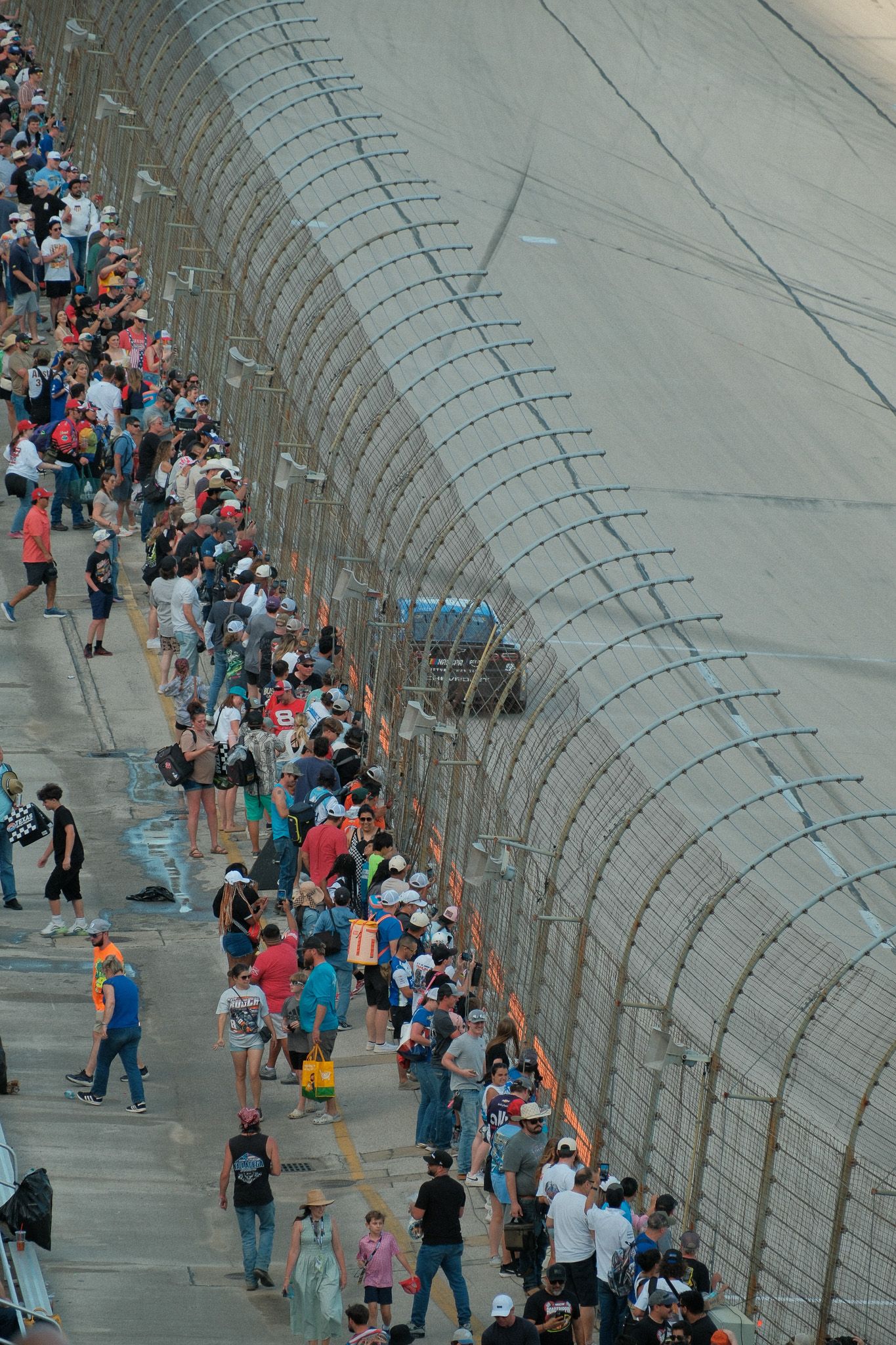 Large crowd of NASCAR fans walking along the track wall behind safety fencing at a speedway racing event.