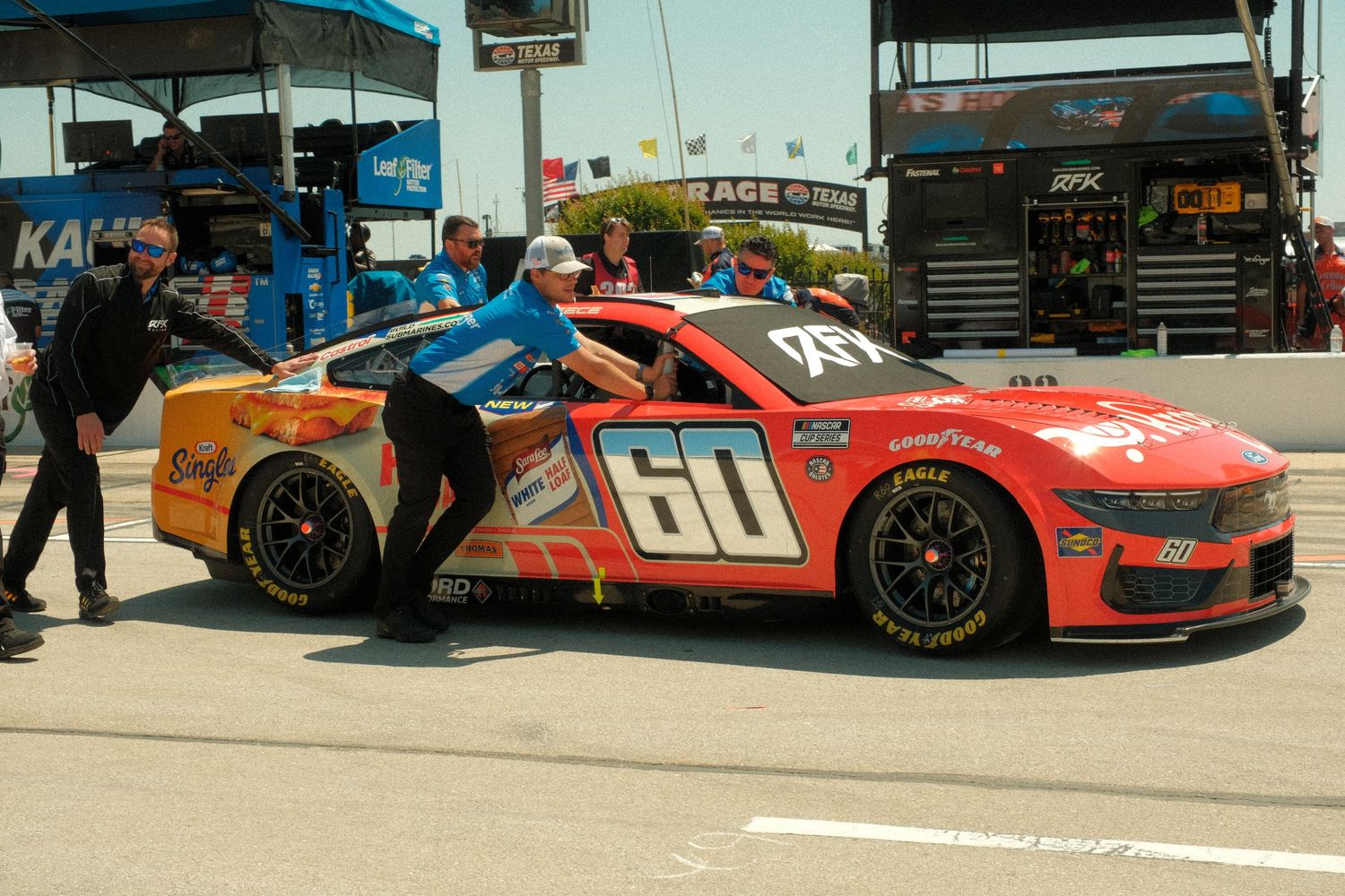NASCAR race car #60 in red livery being pushed through pit lane by crew members at a sunny speedway.