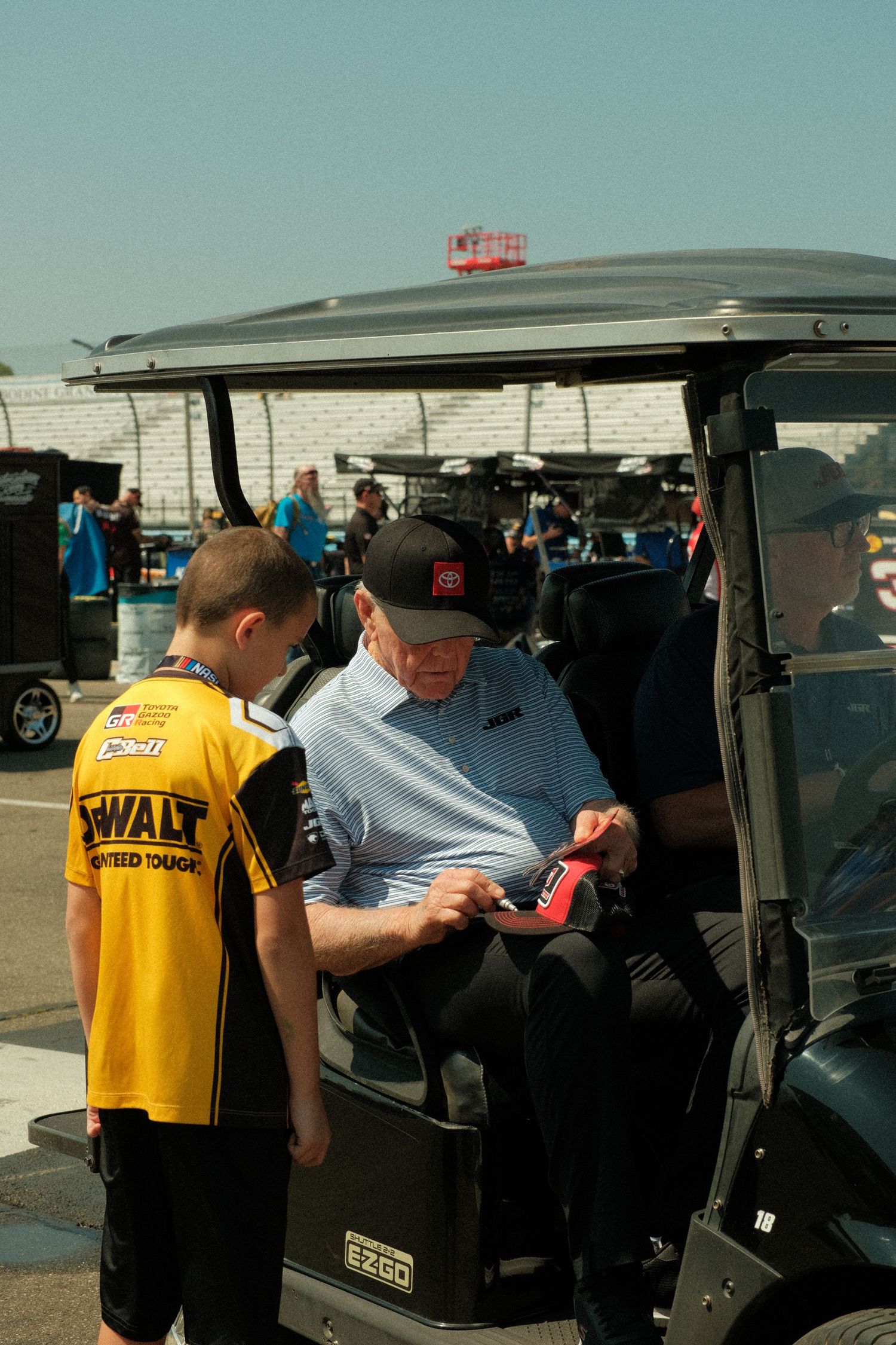 An elderly man Joe Gibbs sits in an EZGO golf cart at a NASCAR event while he signs an autograph for a young fan wearing a DeWalt shirt.