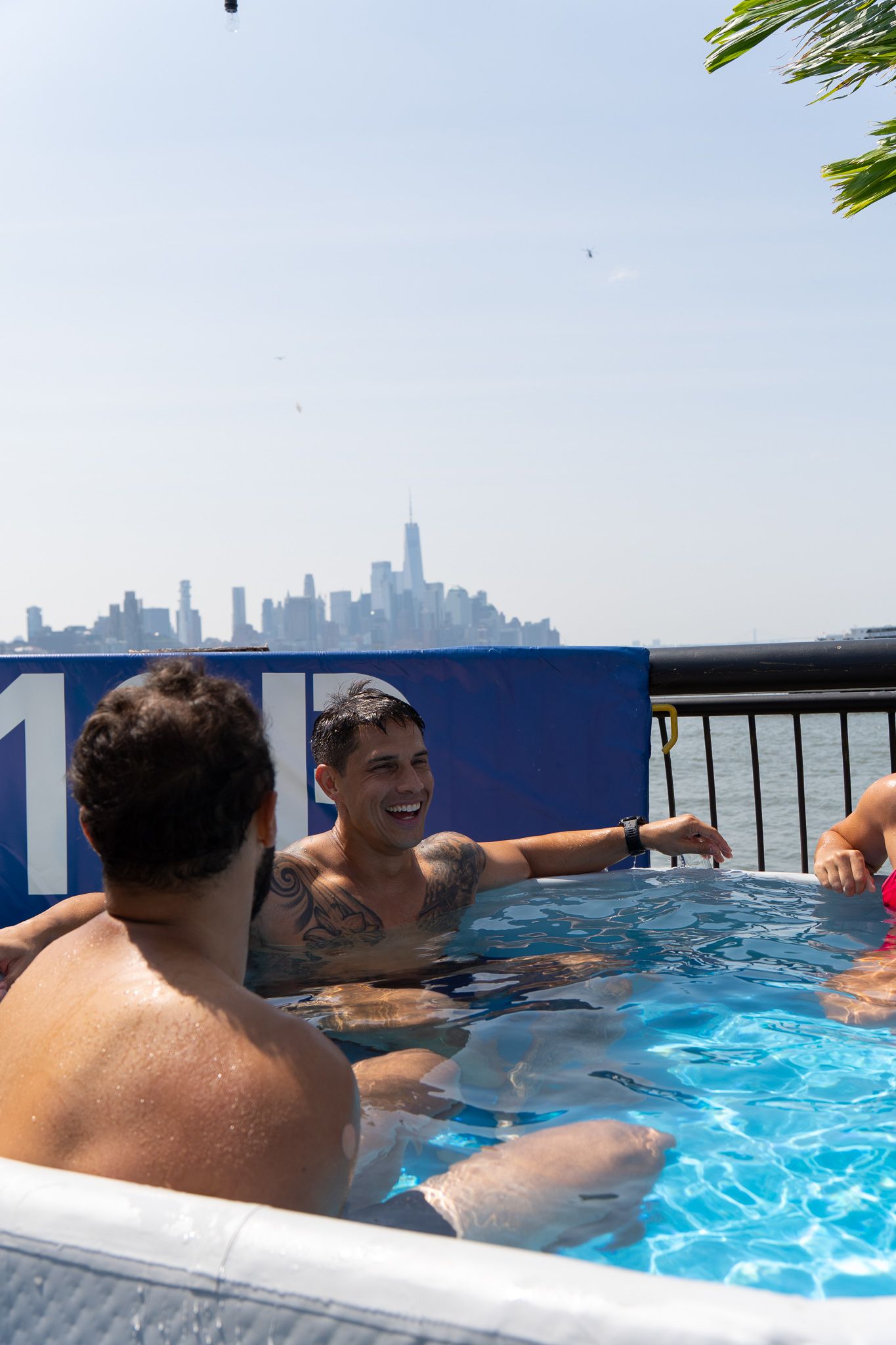 Two tattooed men relaxing in outdoor pool with Manhattan city skyline visible in background.