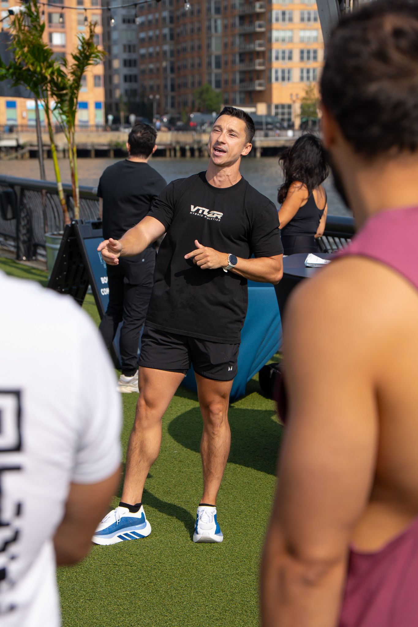 Fit male trainer in black VTLR shirt instructing outdoor fitness class on turf by riverside.