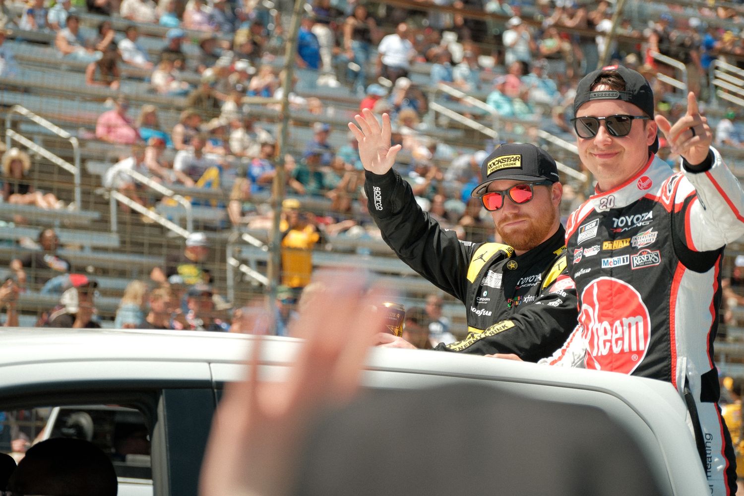 Two NASCAR drivers wave to fans from a parade vehicle at a packed racing event.