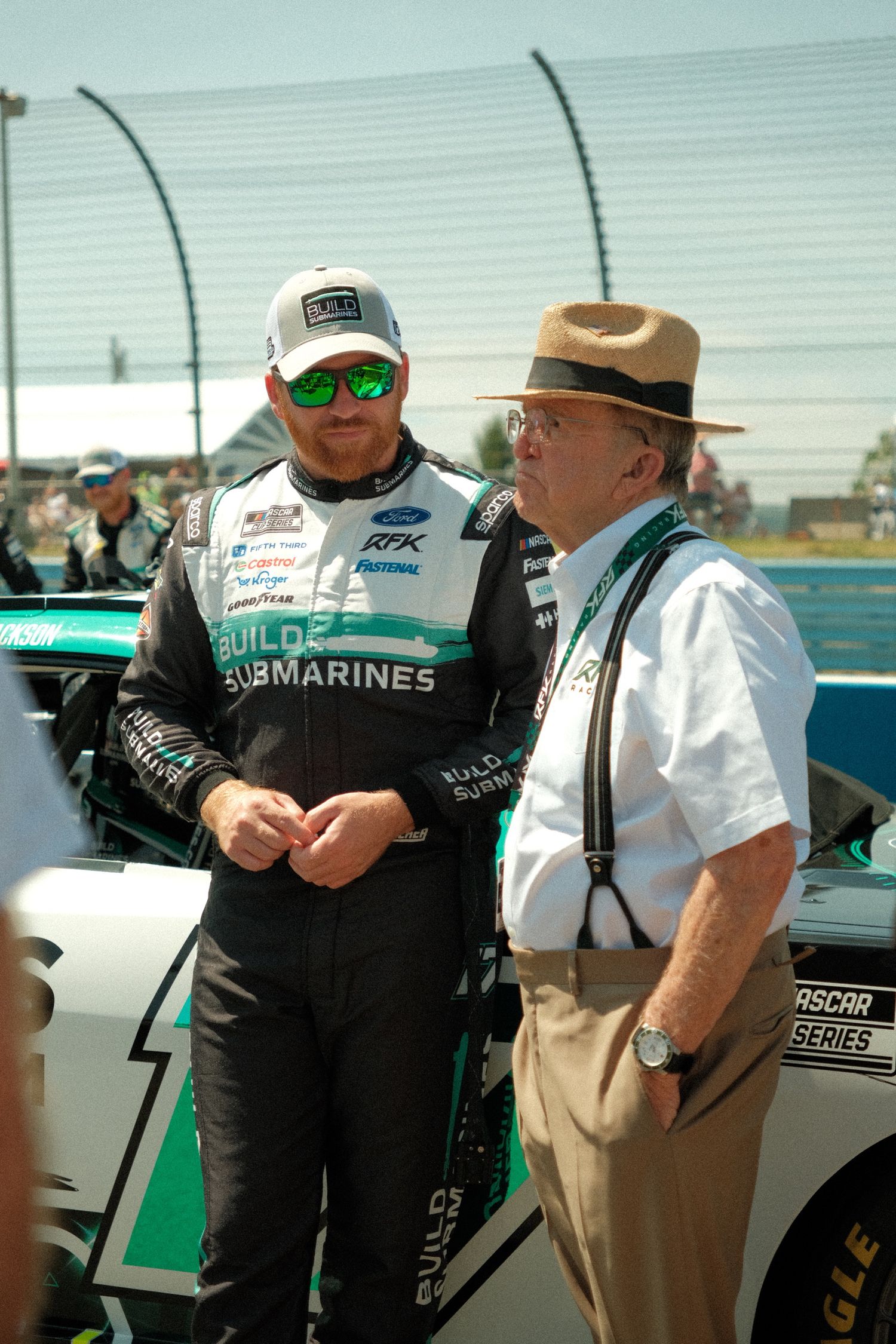 A NASCAR driver Chris Buescher in a Build Submarines uniform talks with an older gentleman Jack Rousch in a tan hat at Watkins Glen.