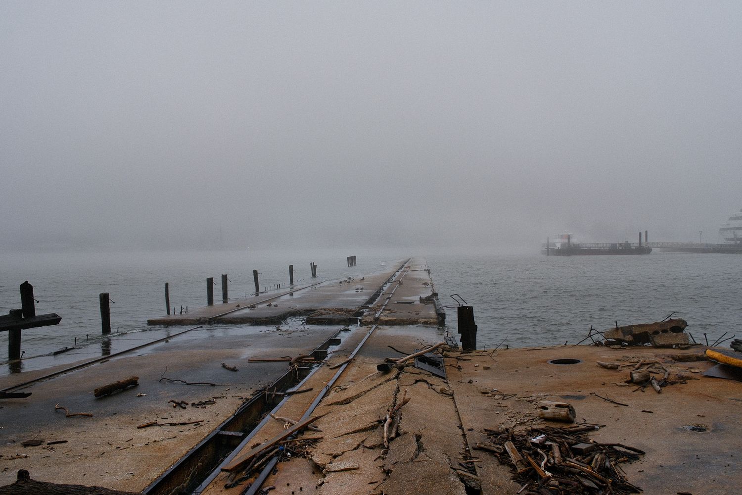 Foggy abandoned wooden pier extending over calm water with debris scattered along shoreline in Hoboken, NJ.