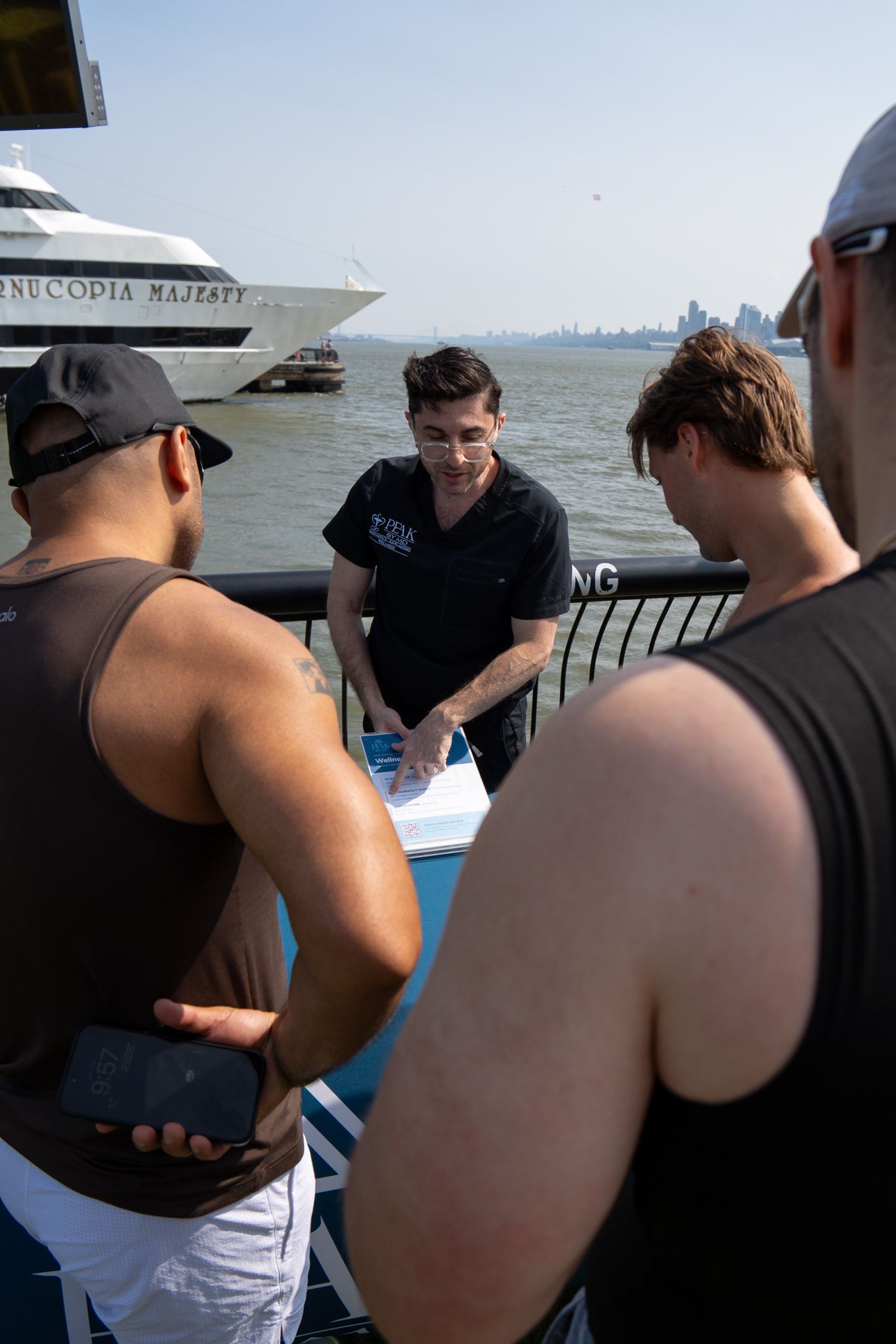 Man checks in guests on a boat dock with NYC skyline and luxury yacht in background on sunny day.