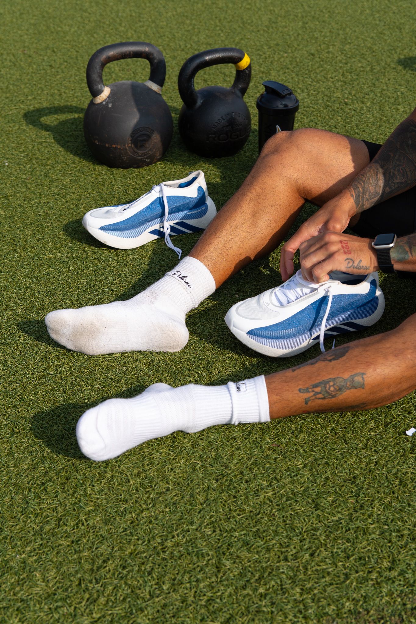 Athlete sitting on turf adjusting blue sneakers next to kettlebells during outdoor fitness training.
