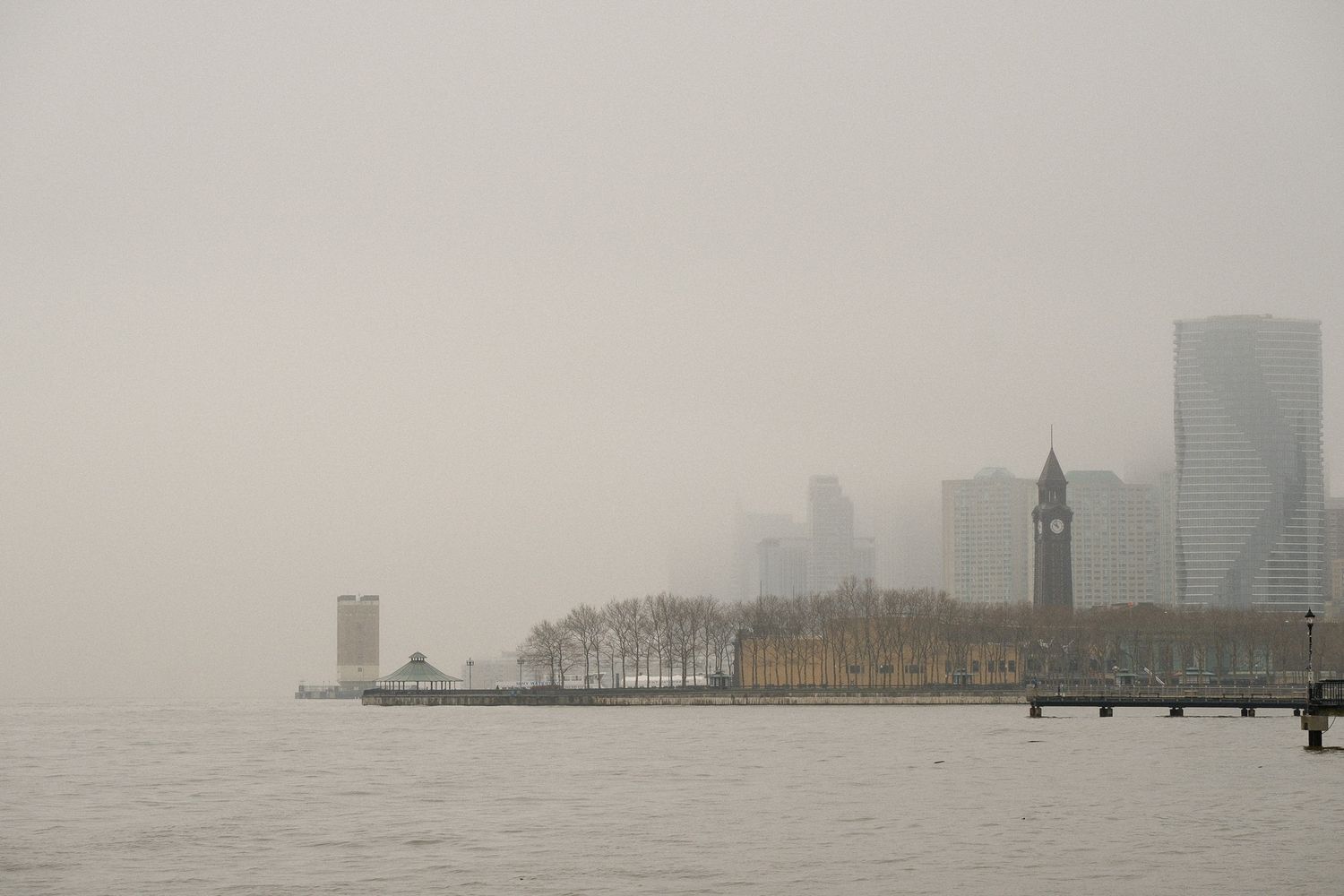 Foggy Hoboken and Jersey City skyline viewed from the water with a Hoboken terminal clock tower and bare trees along the waterfront.