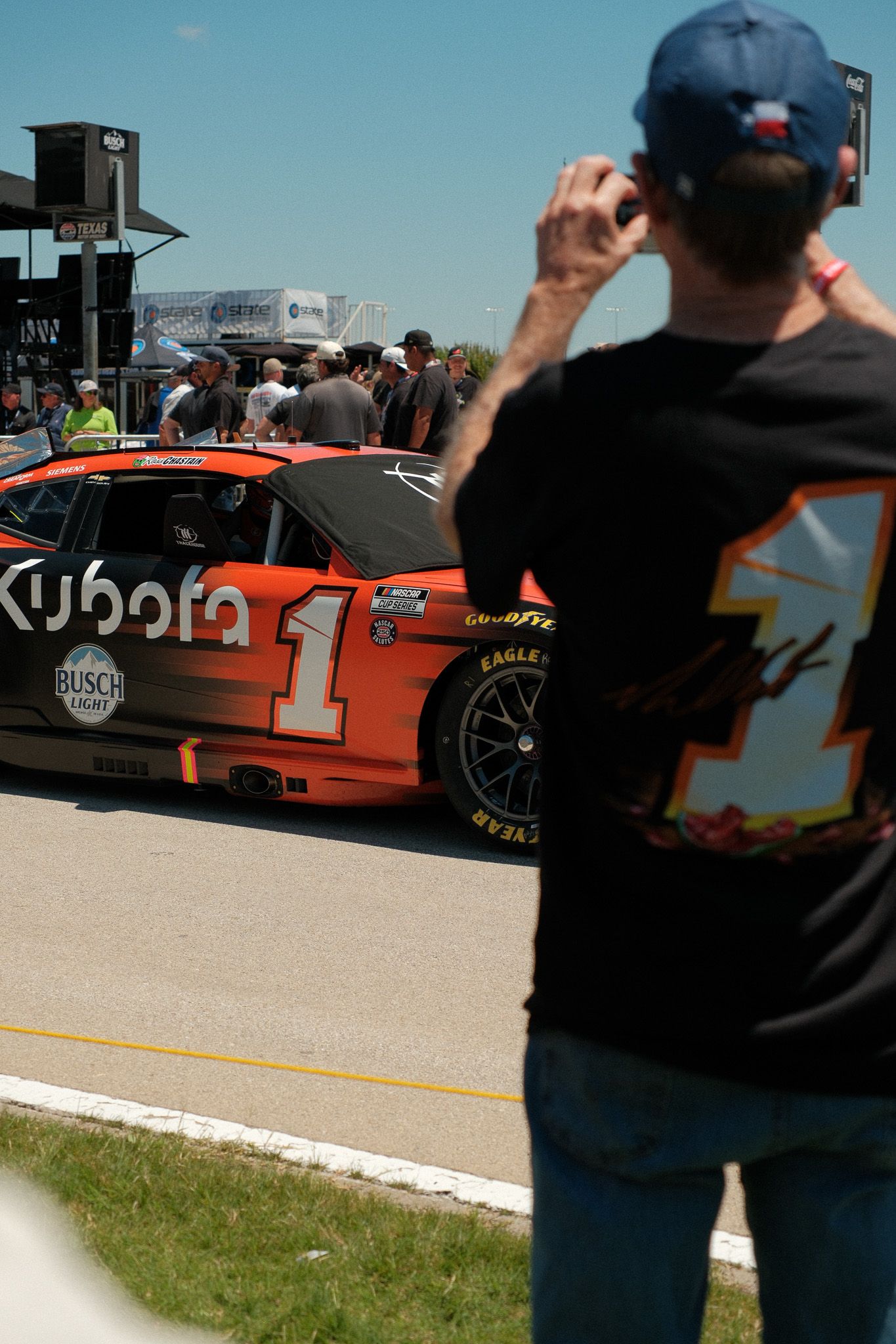 NASCAR fan photographs the Kubota sponsored number 1 race car in the pit area at a sunny racing event.