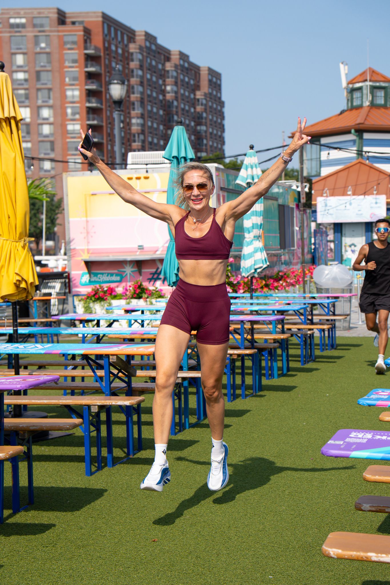 Woman in burgundy workout set jumps joyfully outdoors at a colorful urban fitness event on a sunny day.