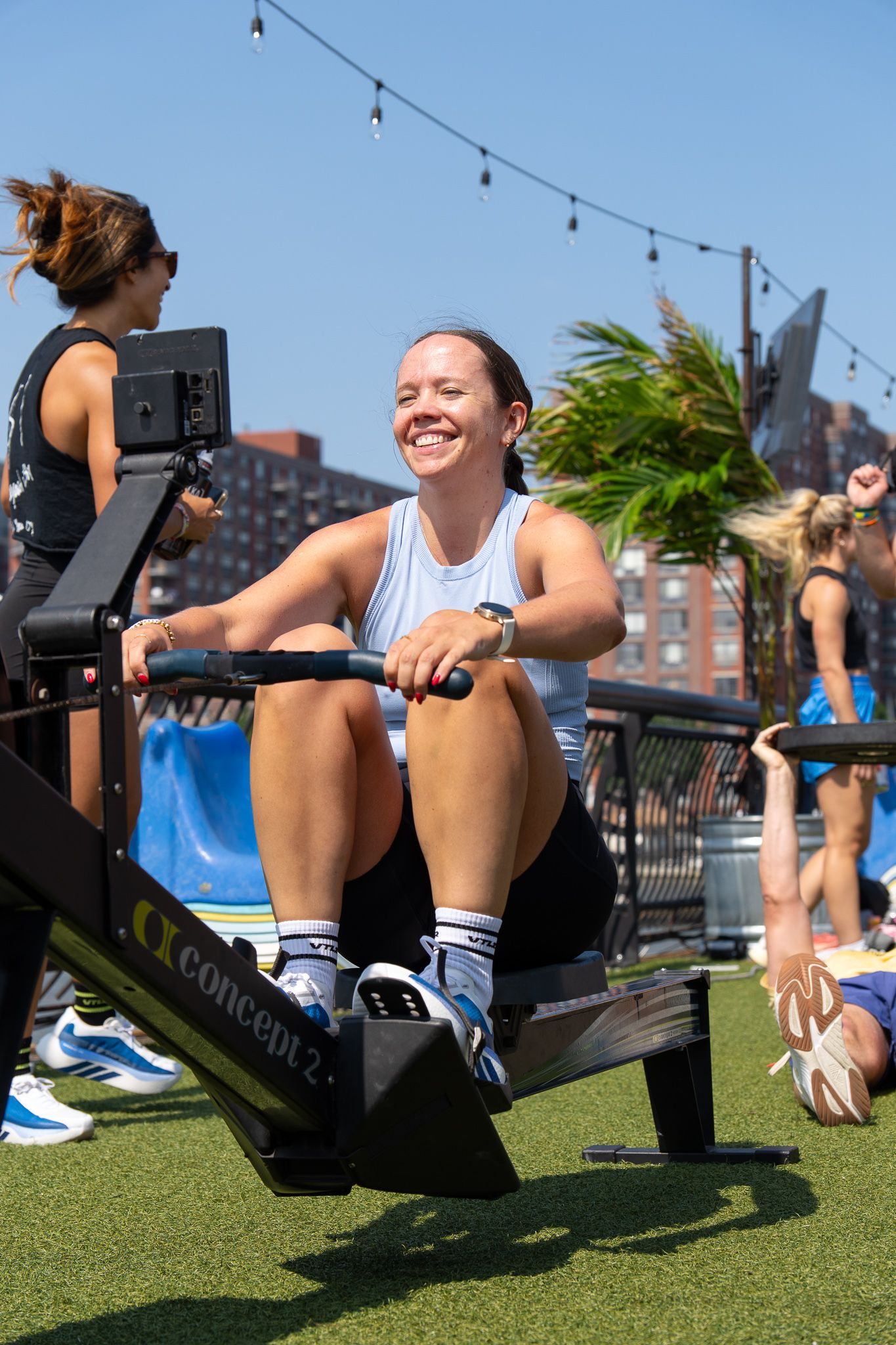 Happy woman smiling while using Concept2 rowing machine during outdoor fitness class on sunny day.