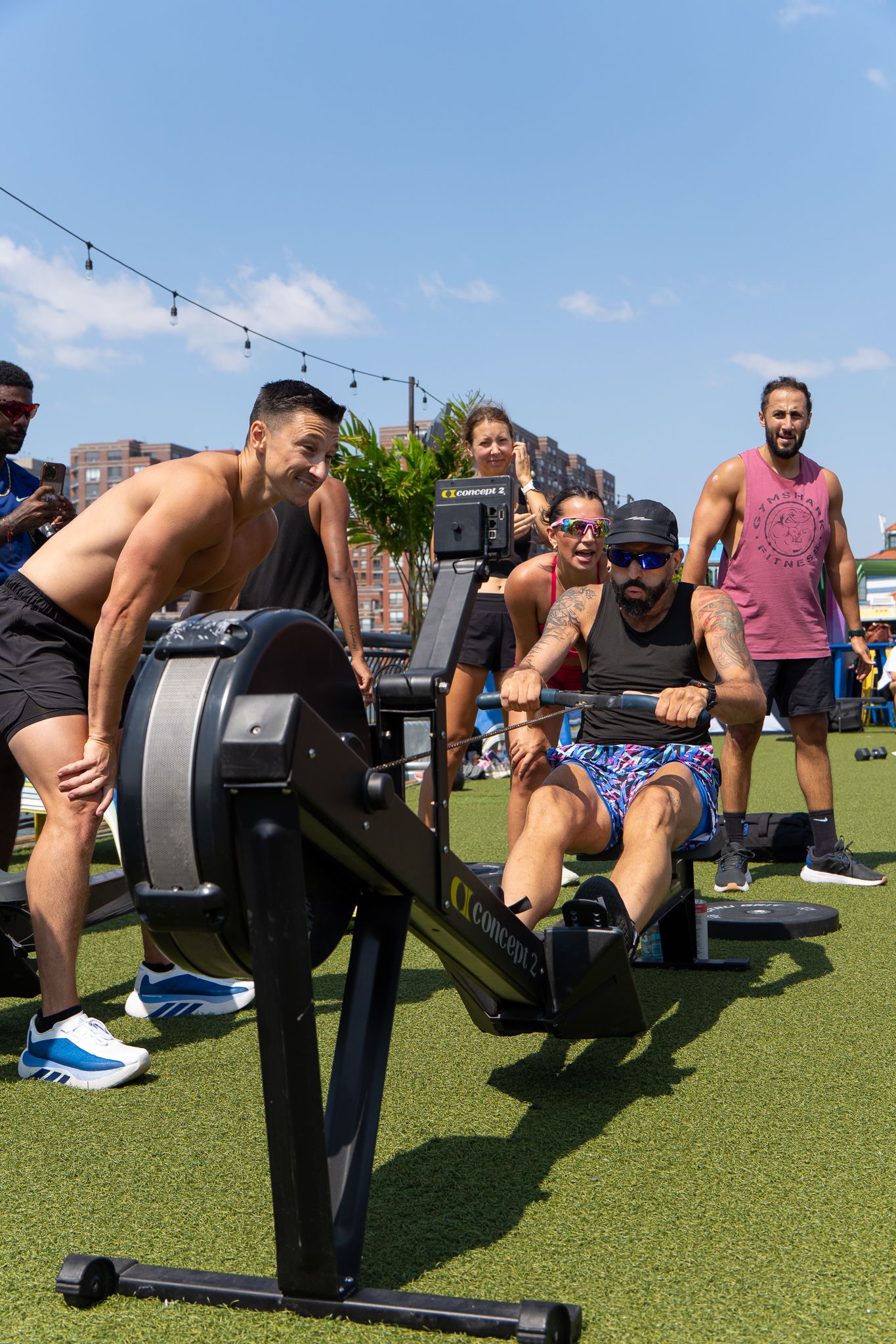 Men competing on rowing machines outdoors at a fitness event on artificial turf under a sunny blue sky.