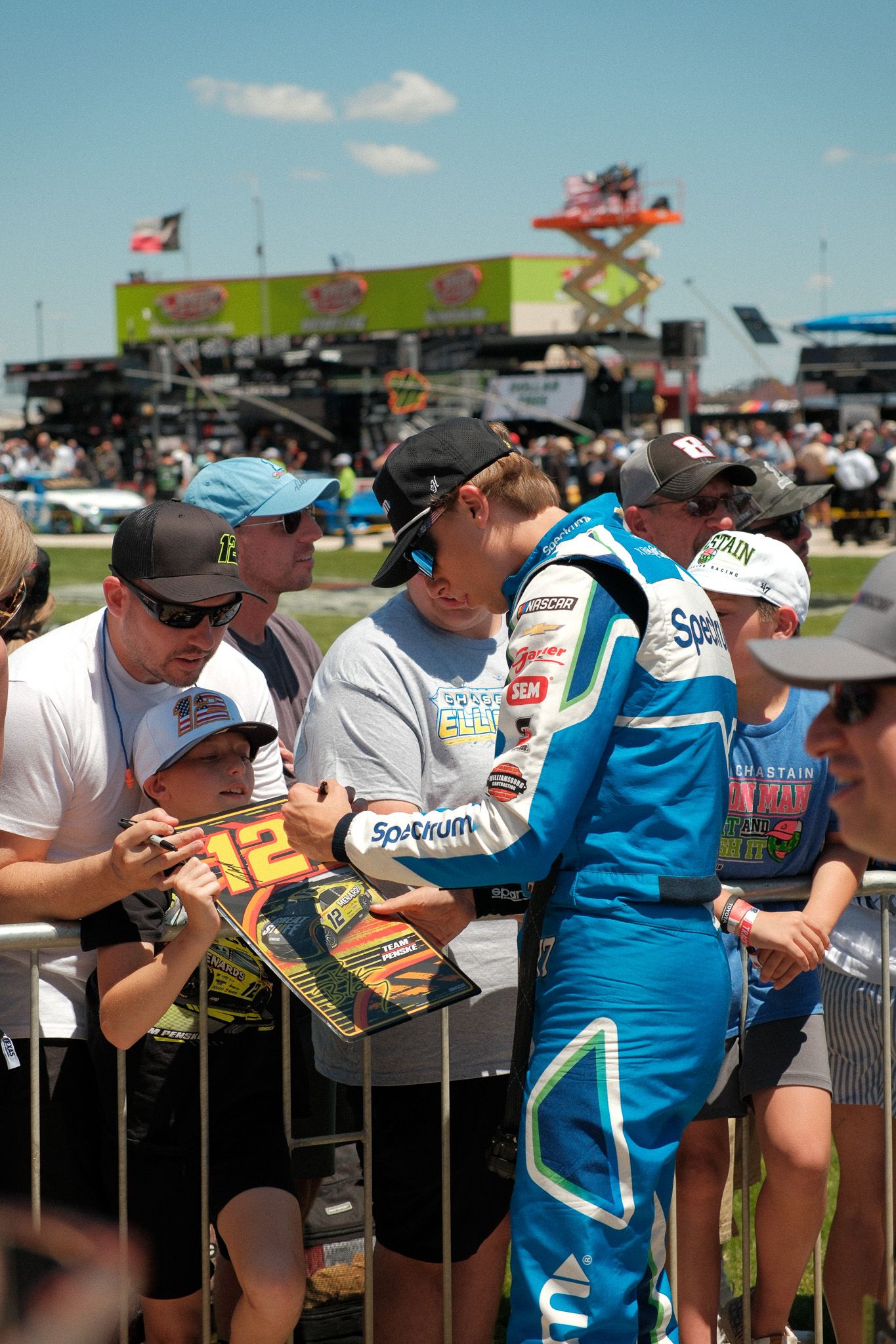 NASCAR driver in Spectrum racing suit signs autographs for fans including a young child at a race event.