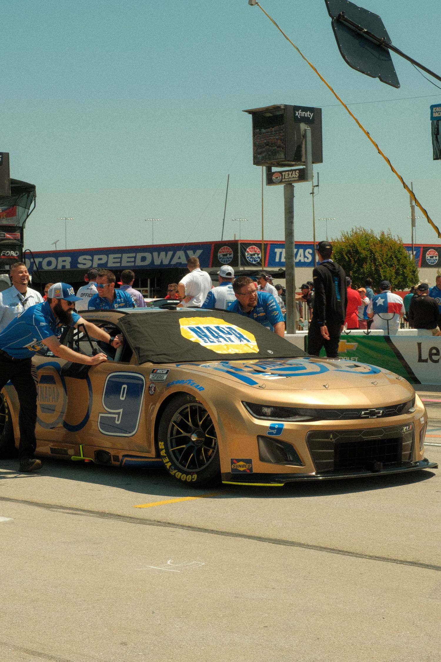 Gold NASCAR #9 NAPA race car being pushed by crew members in pit lane at Texas Motor Speedway.