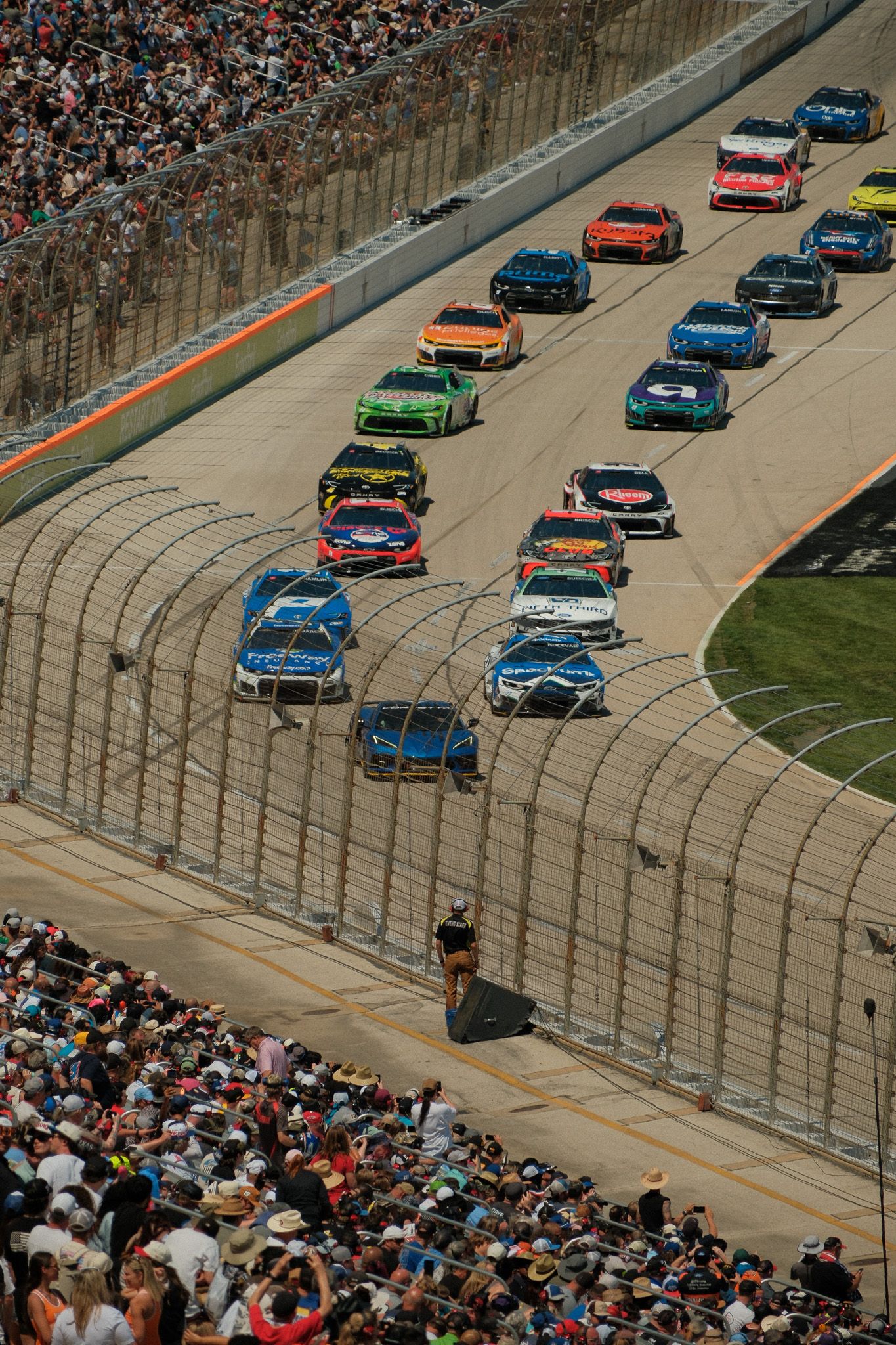 Aerial view of NASCAR stock cars racing on a banked oval track with packed grandstands full of spectators.