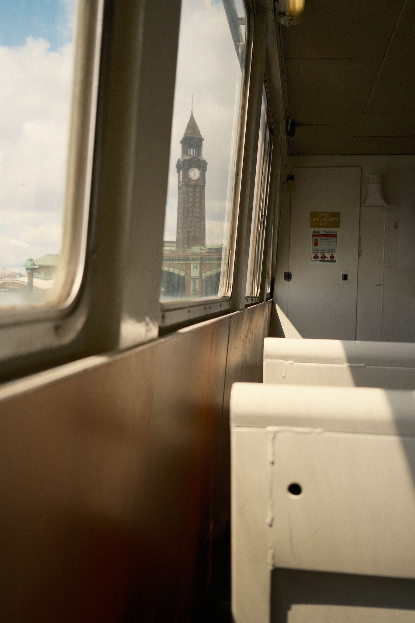 View of a historic clock tower from inside a ferry boat cabin with white seats in Hoboken, NJ.