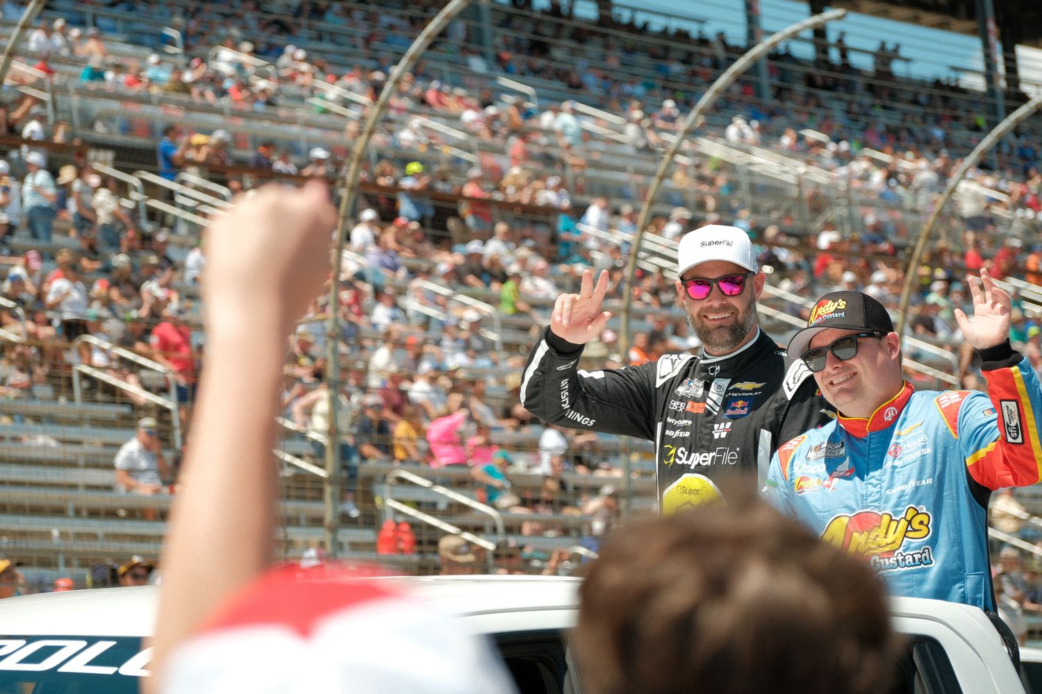 Two NASCAR drivers wave to fans from a vehicle at a packed racing speedway during a sunny race day event.