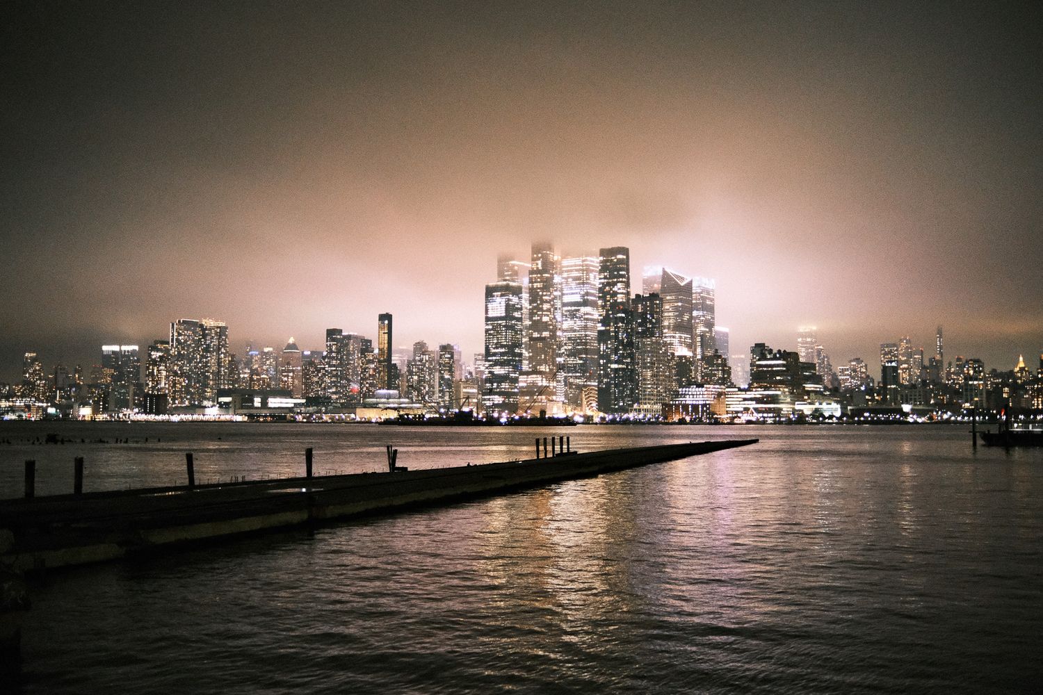 Dramatic nighttime Manhattan skyline reflecting on Hudson River with illuminated skyscrapers through cloudy sky from Hoboken, NJ.