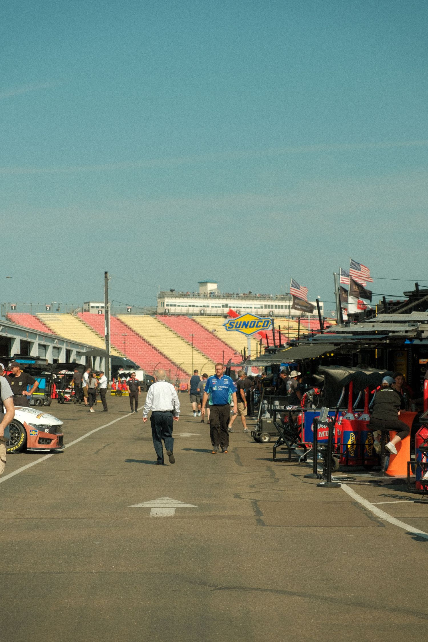 NASCAR pit road with crew members walking near Sunoco branded grandstands under blue sky.
