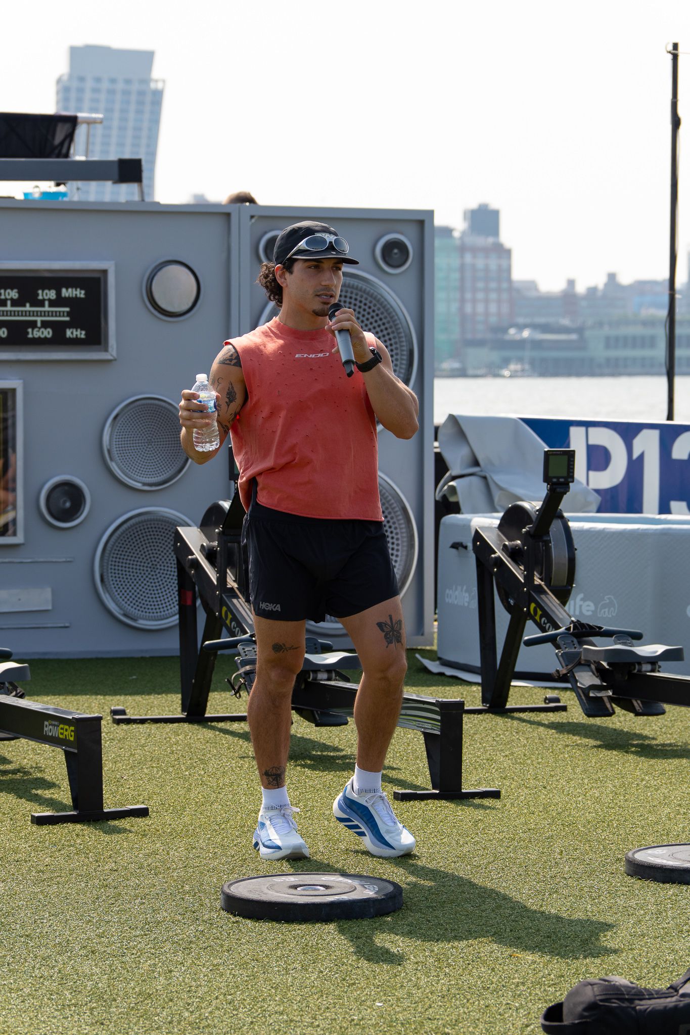 A fitness instructor in a red tank top holds a microphone and water bottle on an outdoor gym turf with rowing machines.