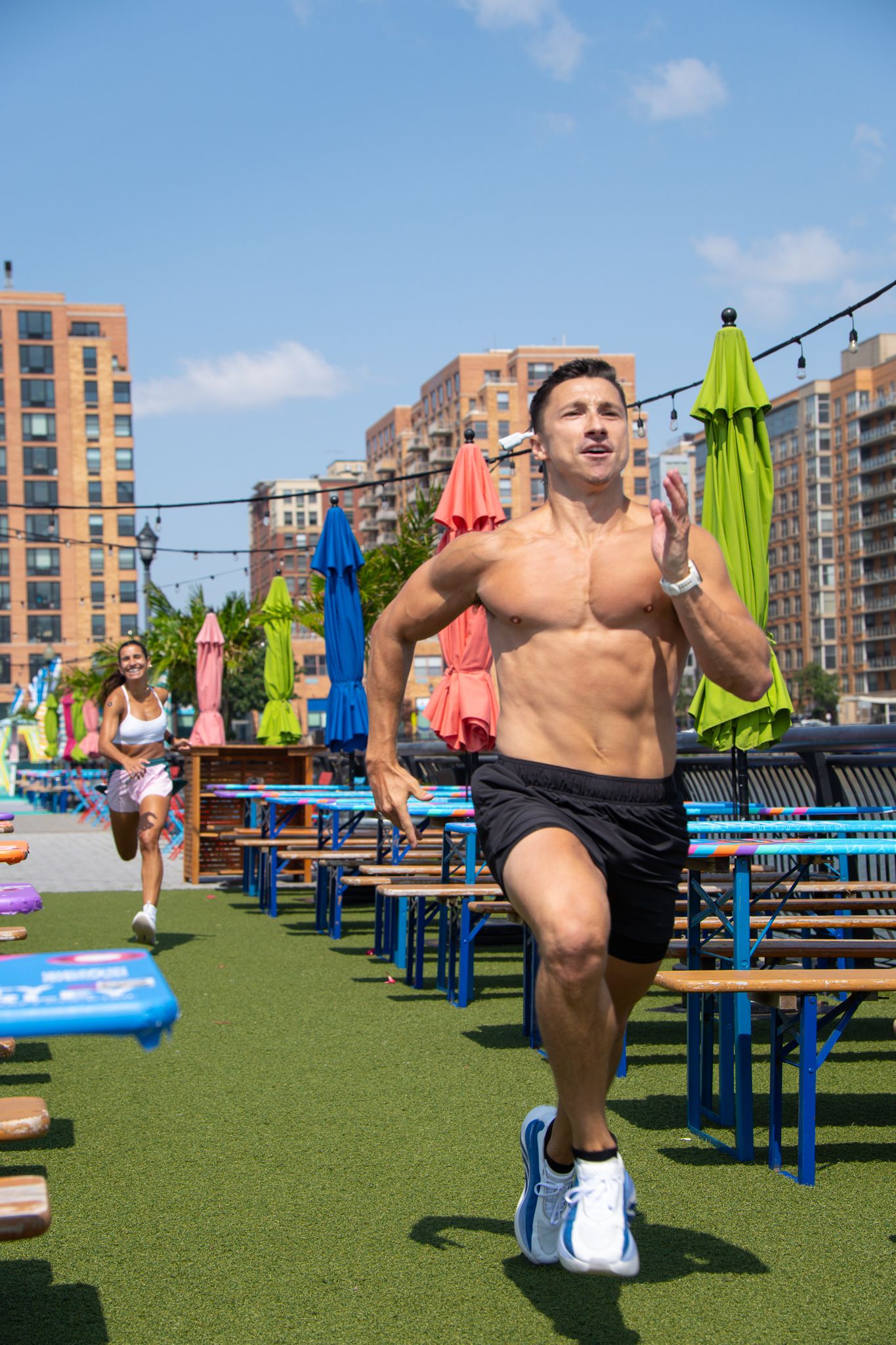 Shirtless athletic man running outdoors at waterfront venue with colorful umbrellas and buildings.