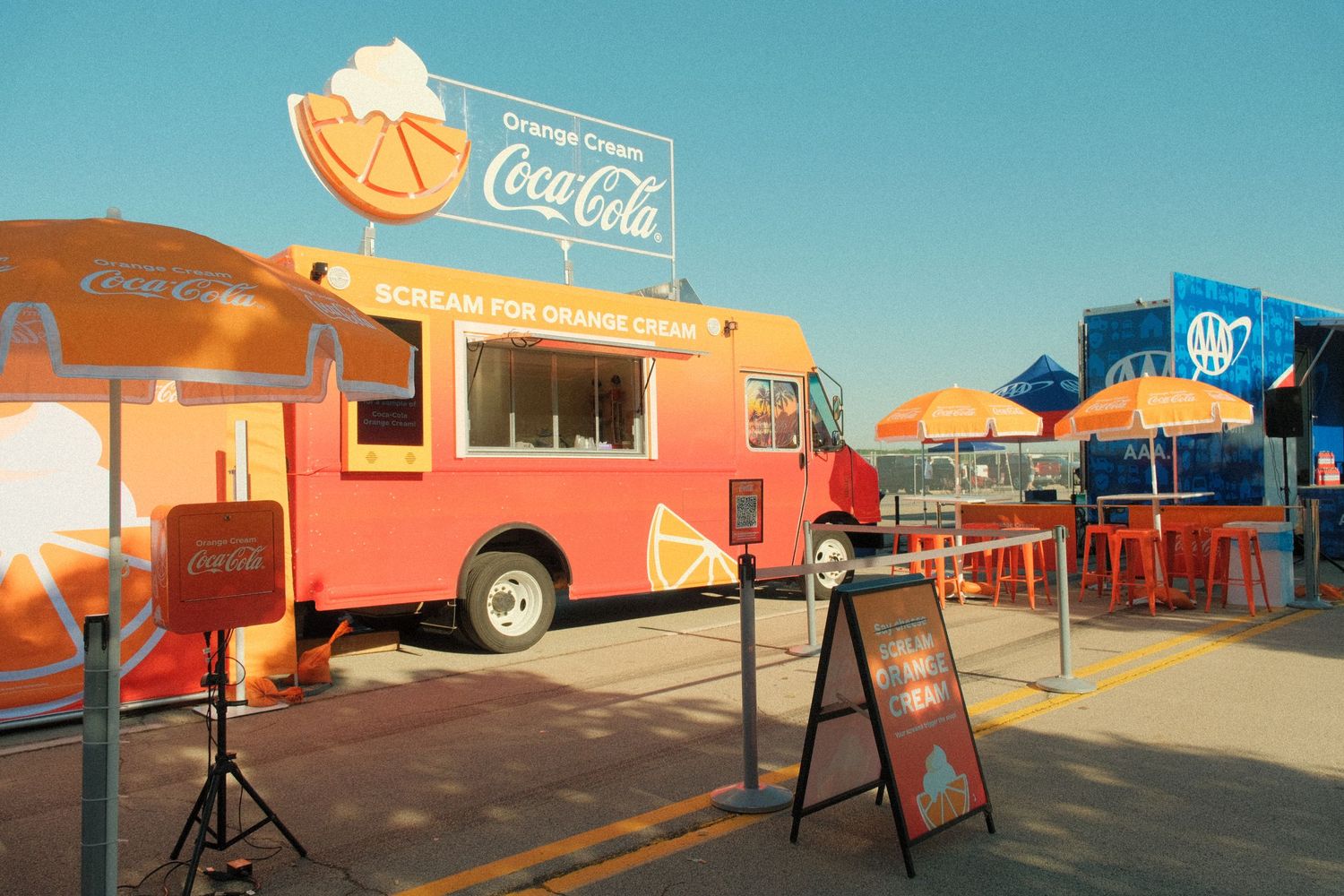 Coca-Cola branded orange cream food truck with colorful umbrellas and outdoor seating at a sunny event.