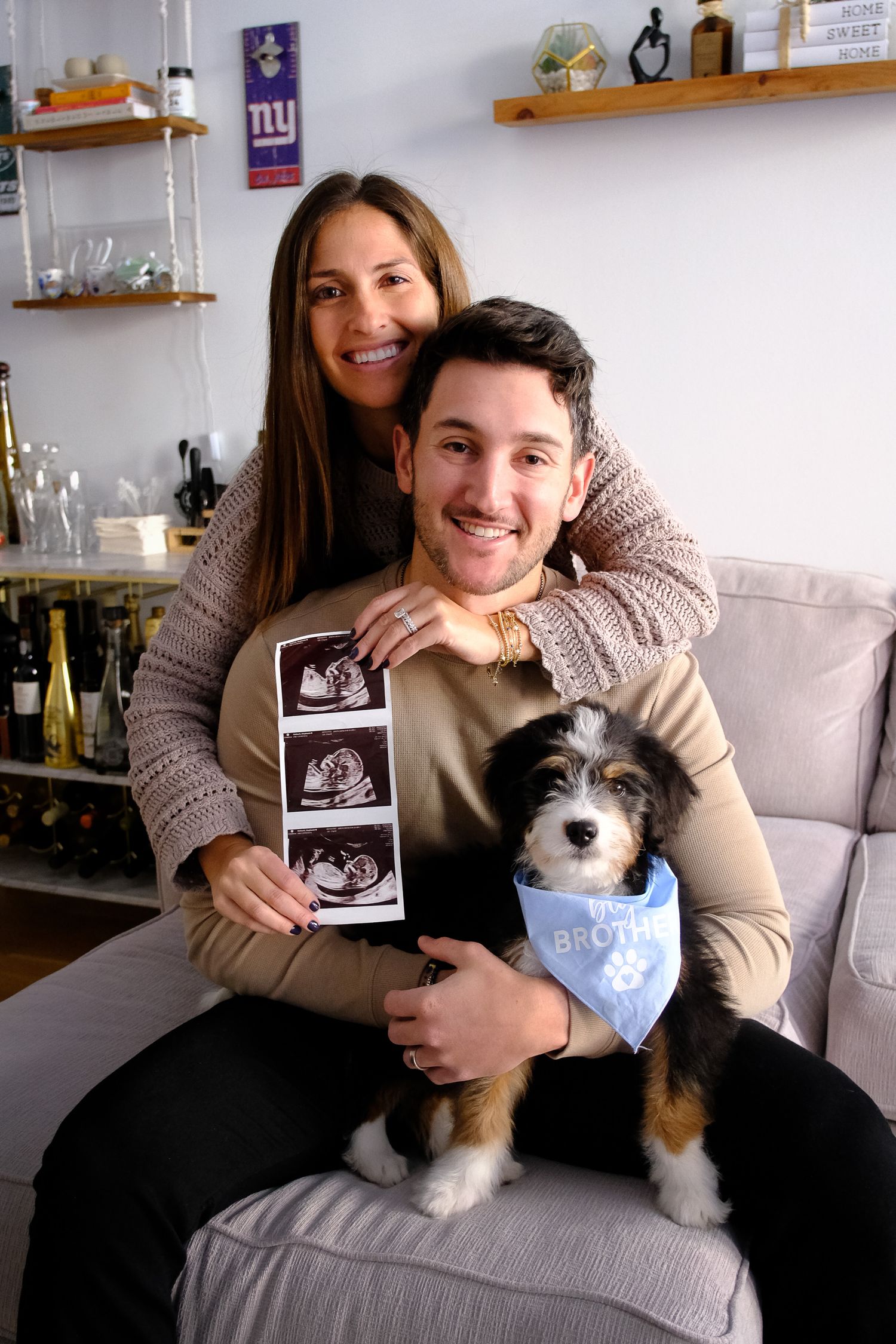 Happy couple holding ultrasound photos with dog wearing Big Brother bandana announcing pregnancy on couch.