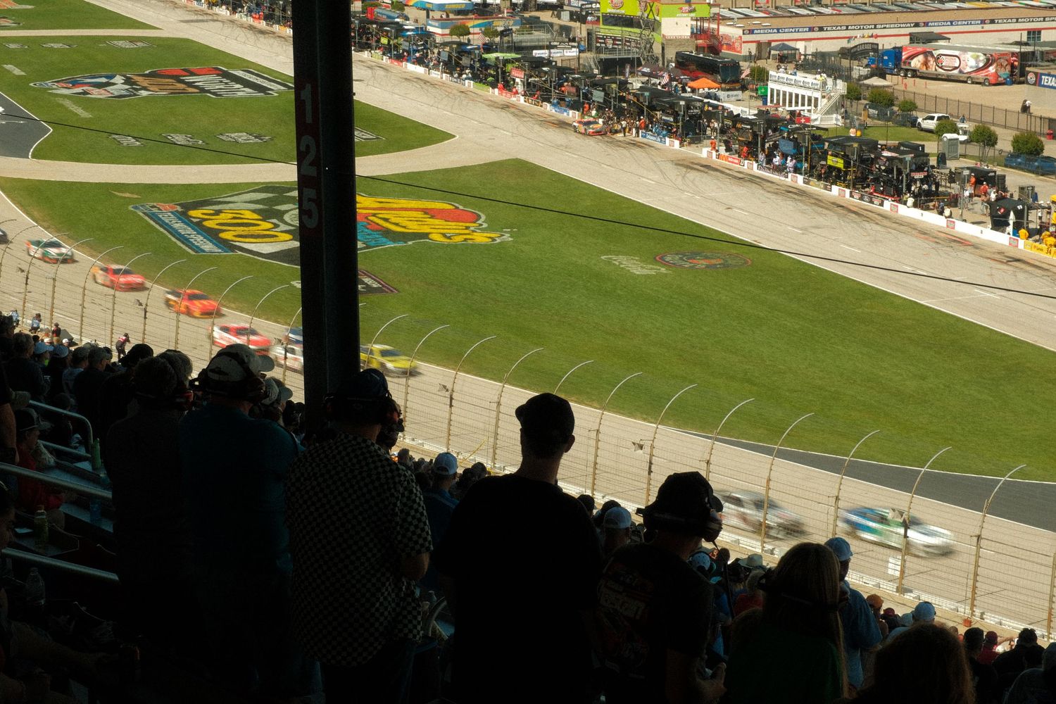 Aerial view of a NASCAR race track with spectators in the foreground watching cars race on the sunlit oval speedway.