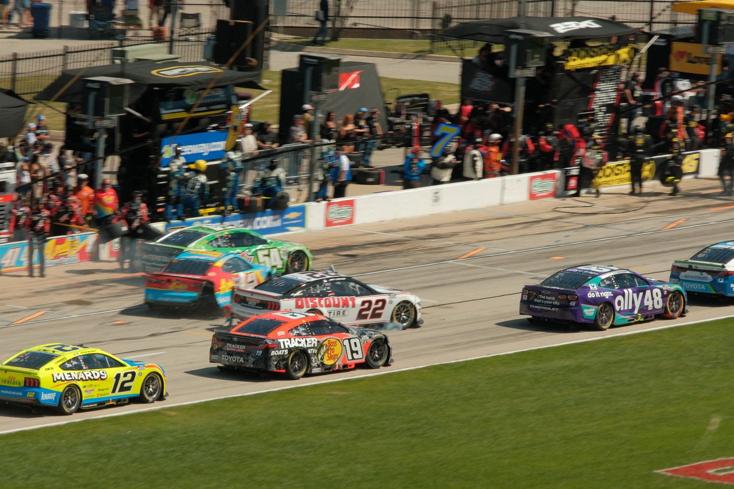 NASCAR race cars numbered 12, 22, and 48 compete on a speedway track with pit crew and fans in the background.