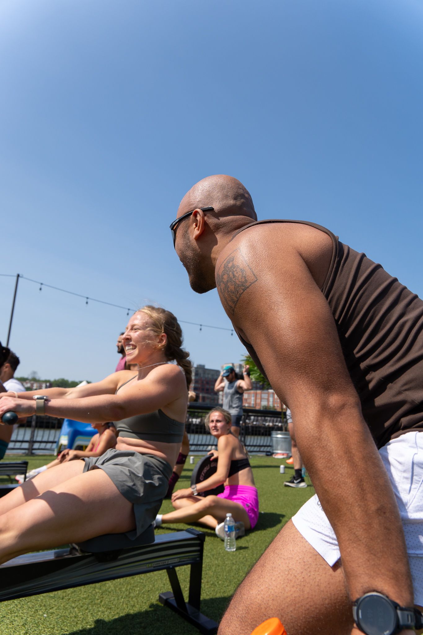 Diverse group enjoying outdoor fitness class on turf, smiling while using exercise benches under clear blue sky.