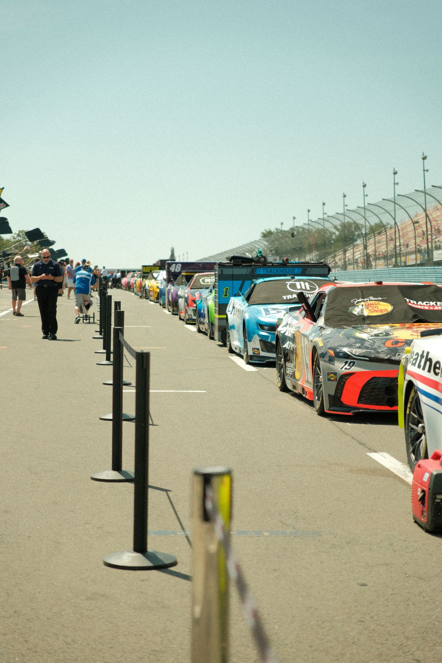 Long row of colorful NASCAR Cup Series race cars lined up on Watkins Glen pit road before a race at a sunny speedway.