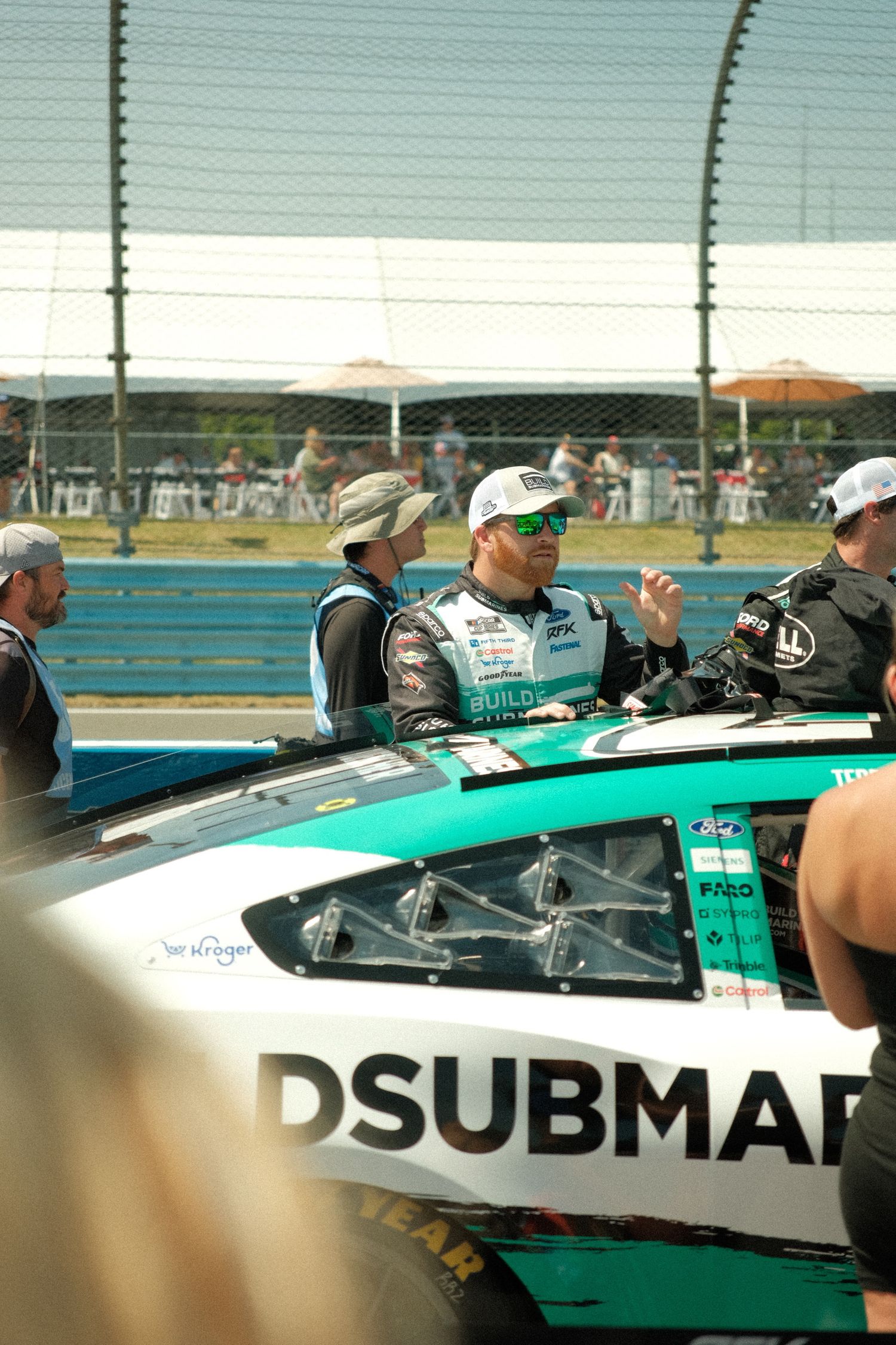 NASCAR driver in teal and white race car with DSUBMAR sponsorship at a racing event on pit road.