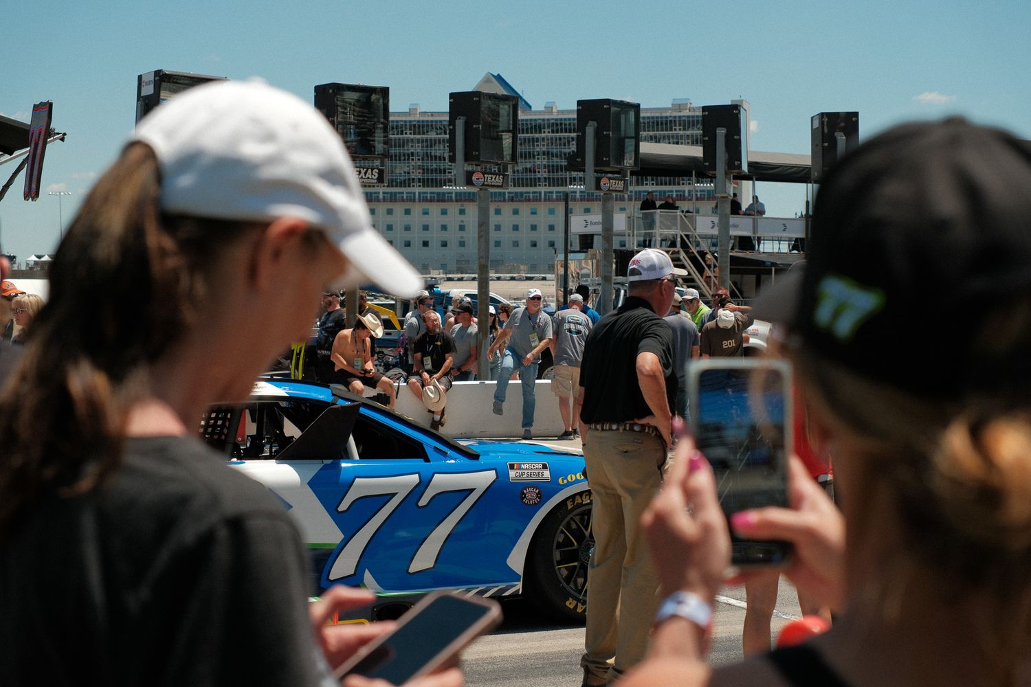 Fans photograph NASCAR car number 77 on display at an outdoor fan event near a waterfront venue.