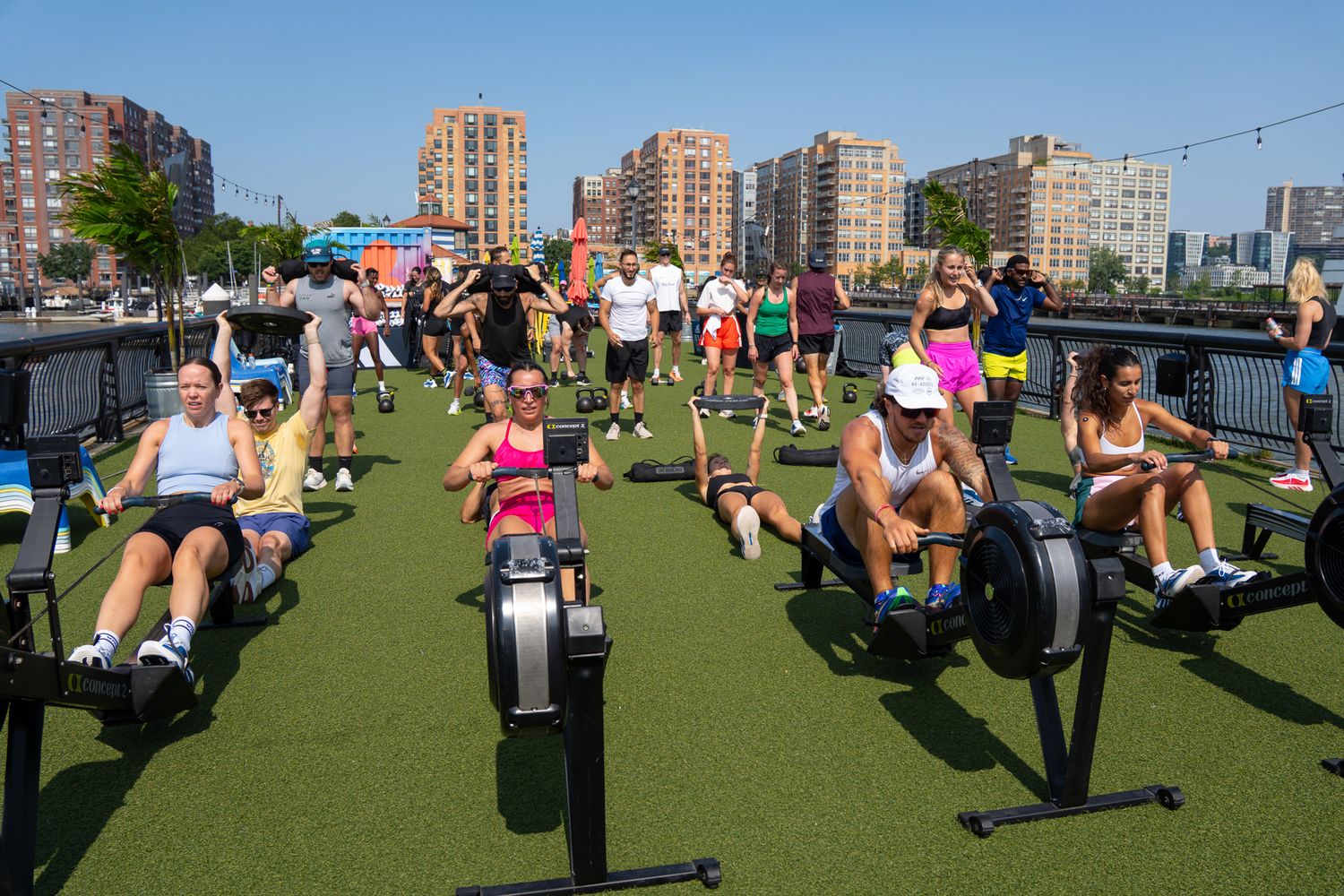 Large group fitness class using Concept2 rowing machines on outdoor turf near waterfront.