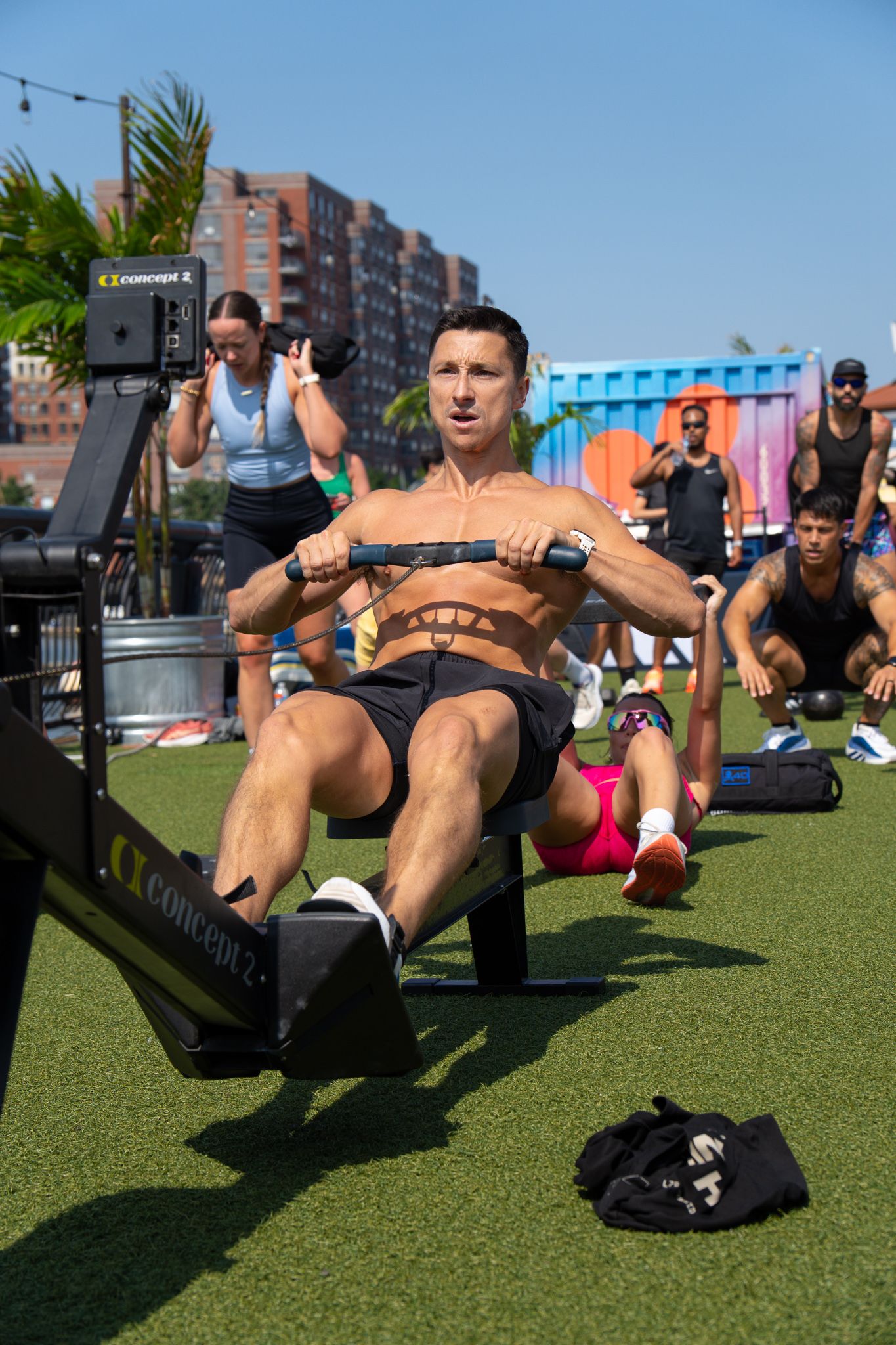 A shirtless man intensely rows on a Concept2 rowing machine during an outdoor group fitness class on artificial turf.