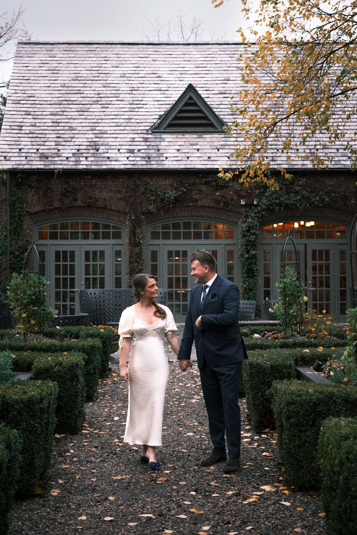 Couple walks hand-in-hand in an autumn garden outside an elegant stone building with green shutters and ivy-covered walls.
