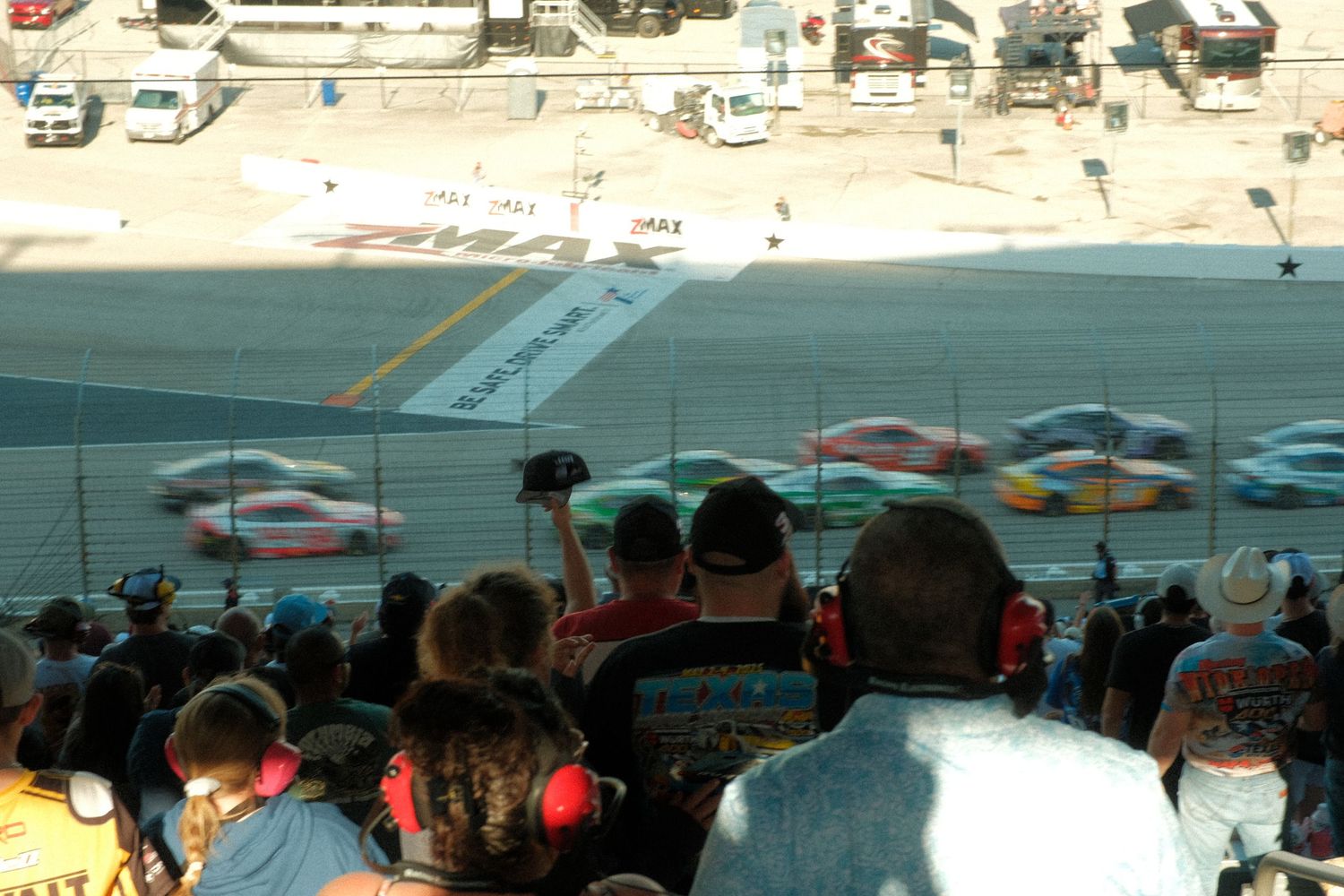 NASCAR race cars speeding past grandstand spectators at a sunny outdoor racing event.