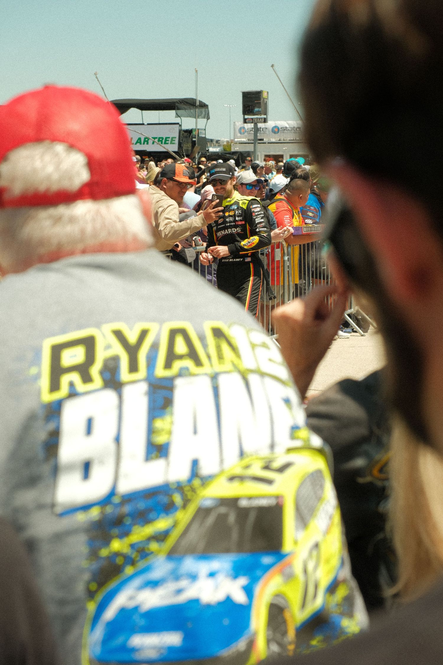 NASCAR fans watch Ryan Blaney interact with crowd at a race event, fan wearing Ryan Blaney shirt in foreground.