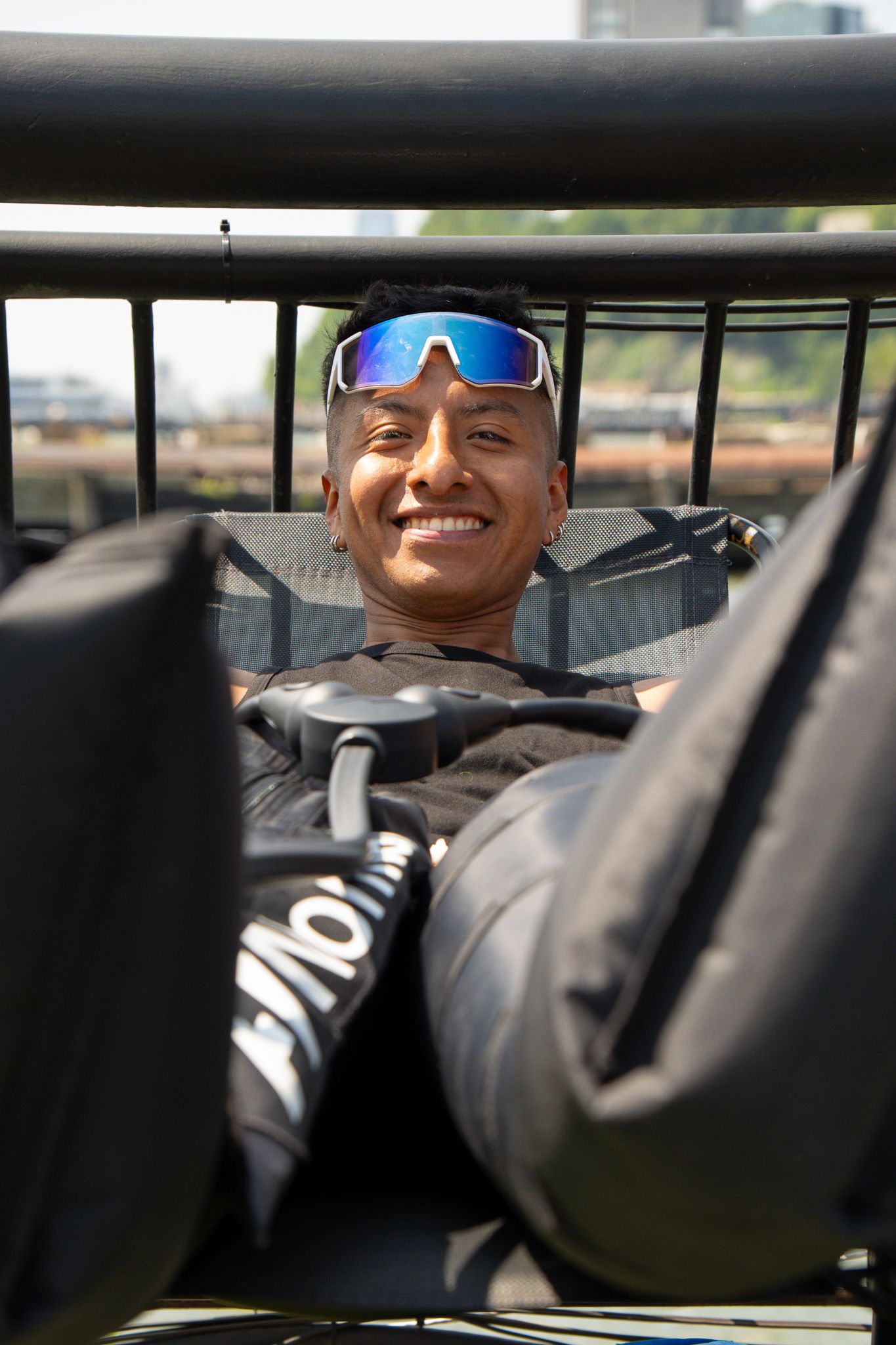 Smiling man with blue mirrored sunglasses resting outdoors with gym bags in foreground.