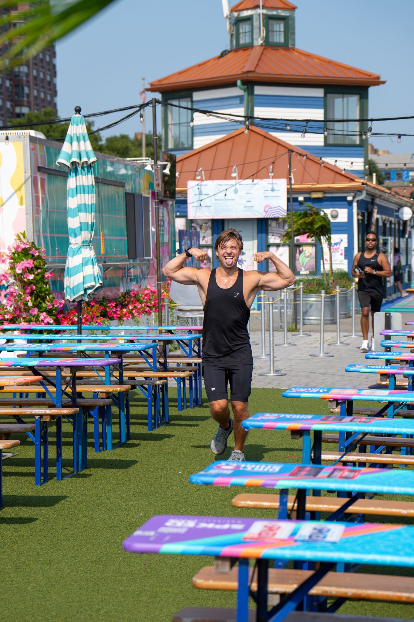 Fit man flexes muscles at colorful outdoor venue with picnic tables on a sunny day.
