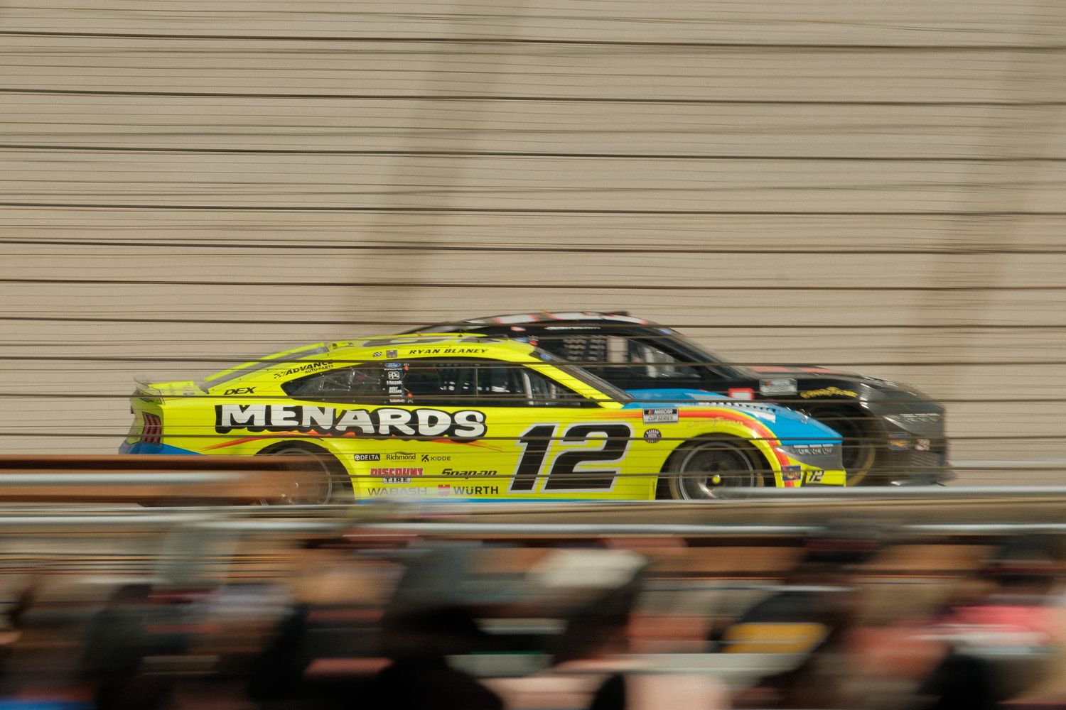 NASCAR race car #12 with Menard's sponsorship speeding on track, motion blur visible on yellow and black vehicle.