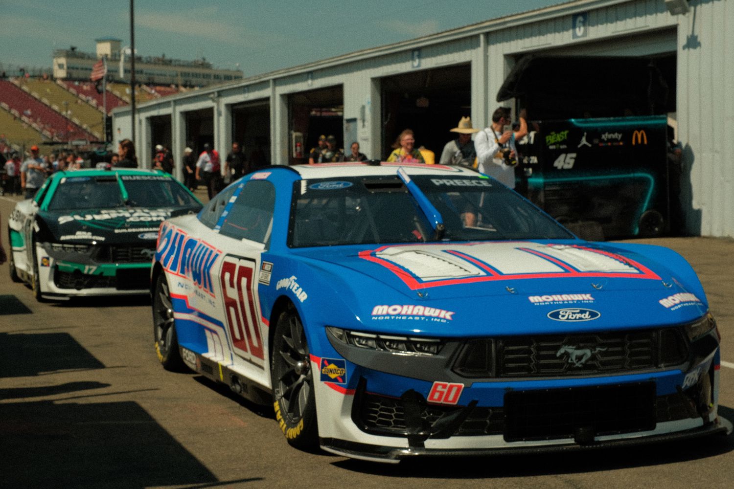 Blue and red Ford Mustang NASCAR number 60 lines up in pit lane before a race at a speedway.