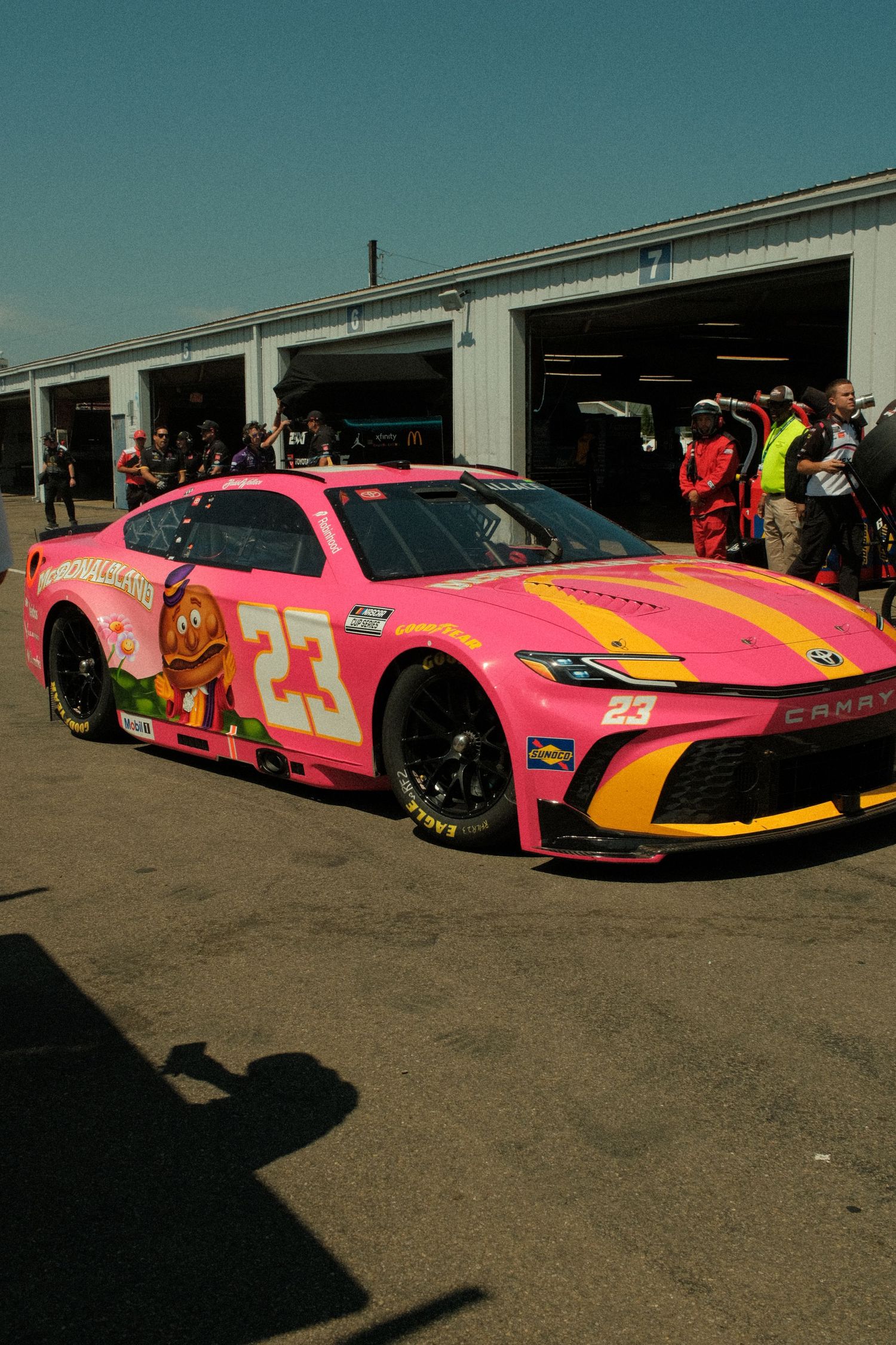 Pink NASCAR #23 Toyota Camry race car with colorful graphics in pit lane garage area on sunny day.