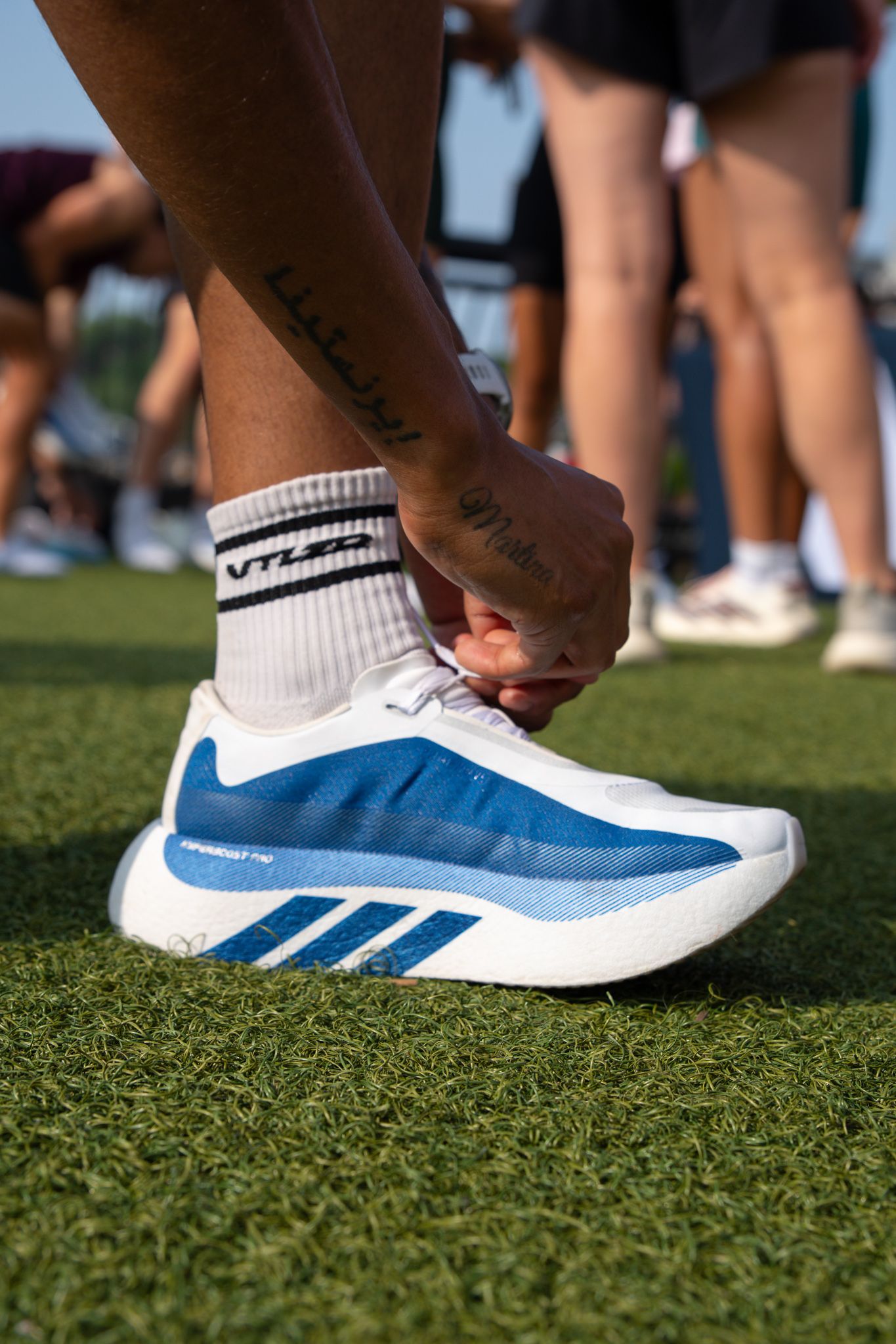 Person tying blue and white Adidas running shoes on green turf while wearing VTL crew socks.