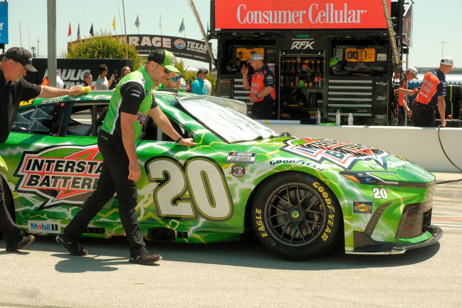 NASCAR #20 Interstate Batteries green car being pushed by crew members in pit lane at a Consumer Cellular sponsored race.
