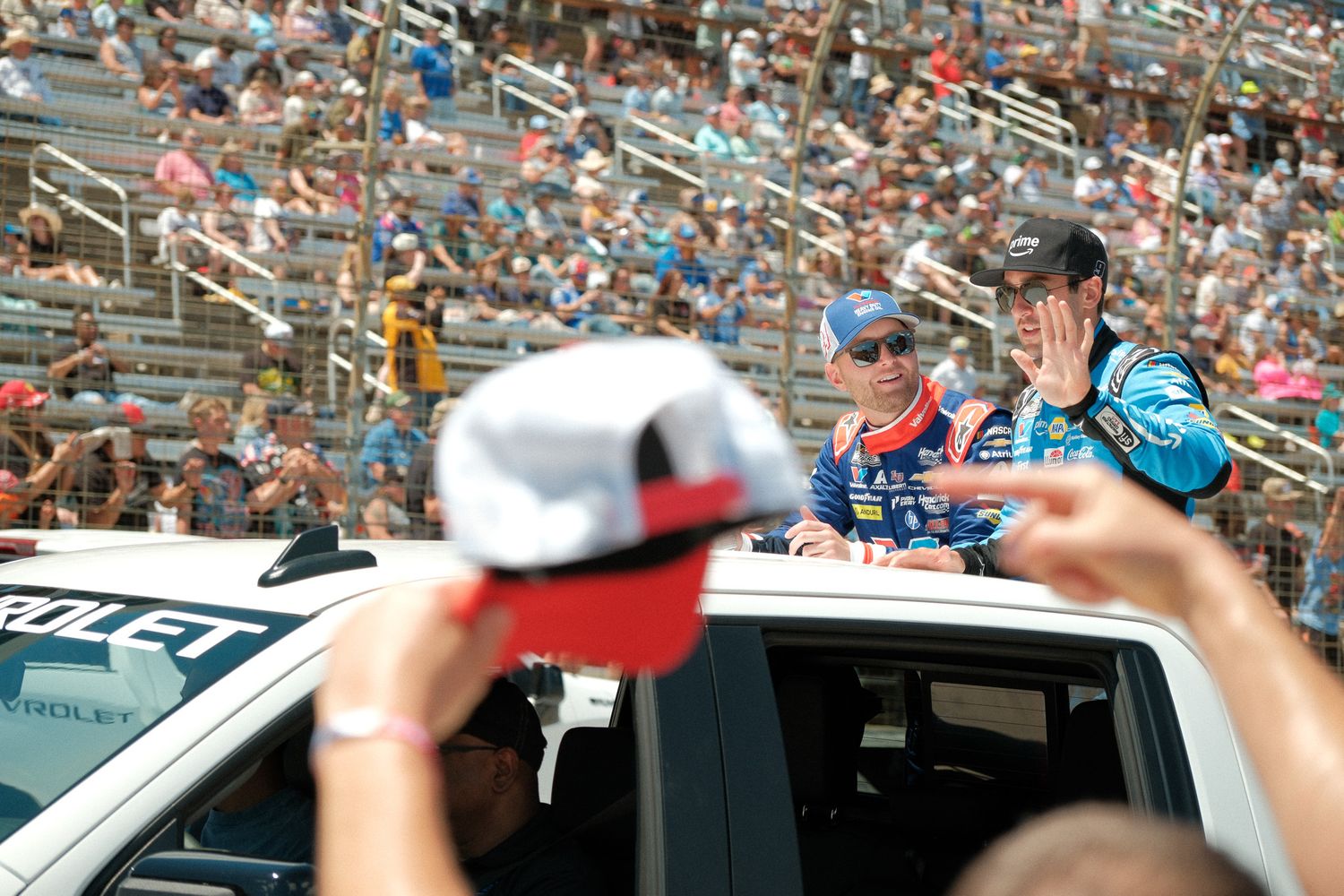 NASCAR drivers wave to fans from a truck during a pre-race parade at a packed speedway on a sunny day.