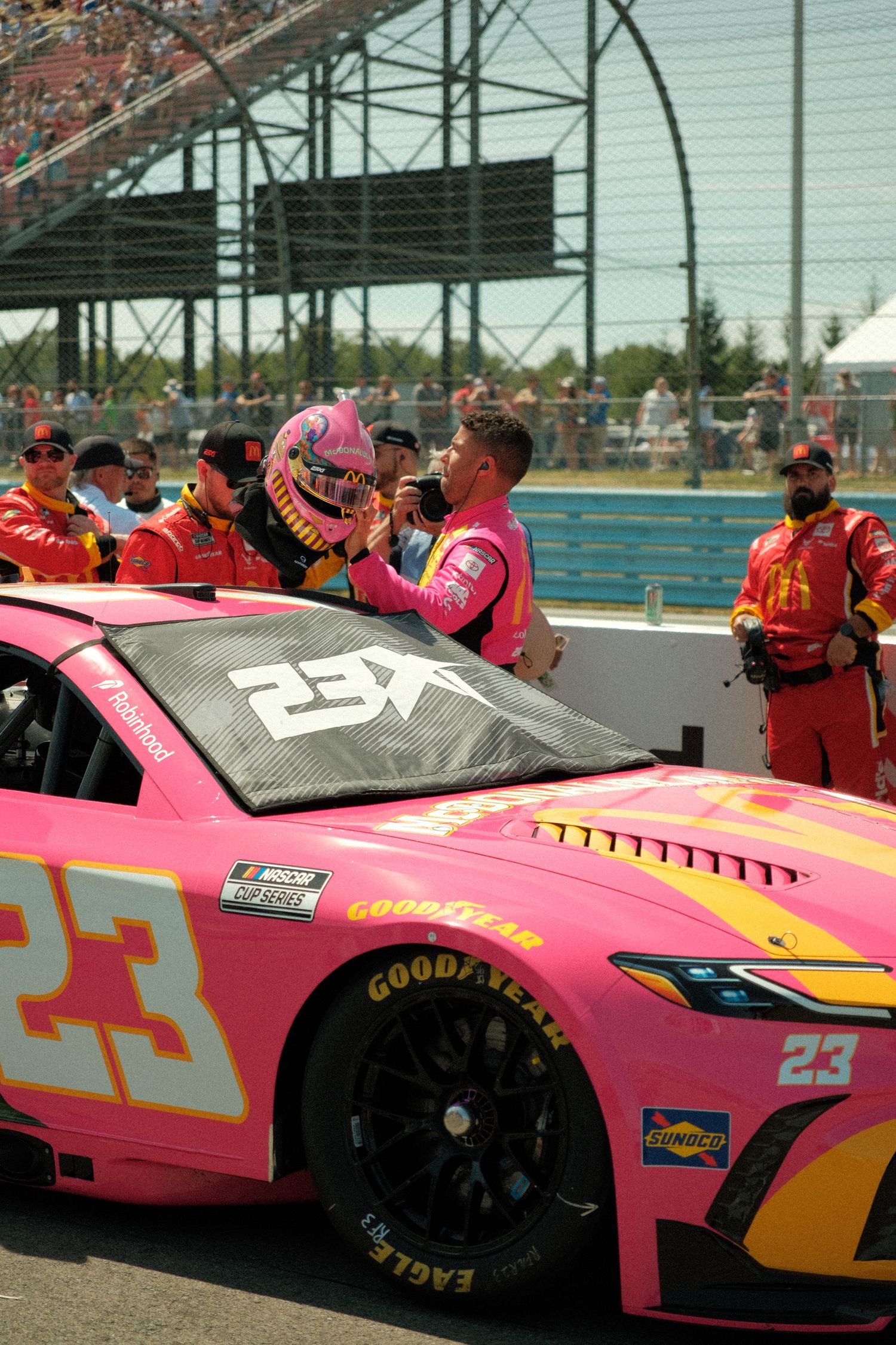 A NASCAR Bubba Wallace driver in a pink helmet stands beside the pink number 23 Toyota before a race at Watkins Glen.